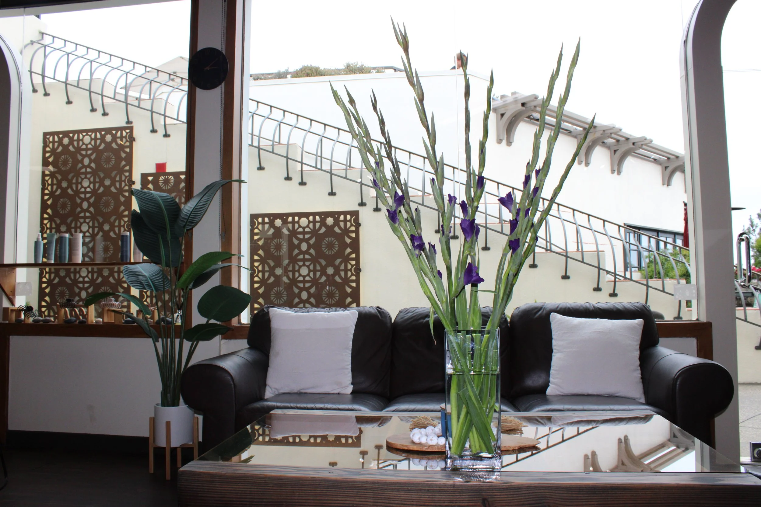 Interior of a cafe with a large glass vase of tall purple and green flowers on a reflective table, a black leather sofa with white pillows, an indoor plant, and large windows showing an outdoor staircase.