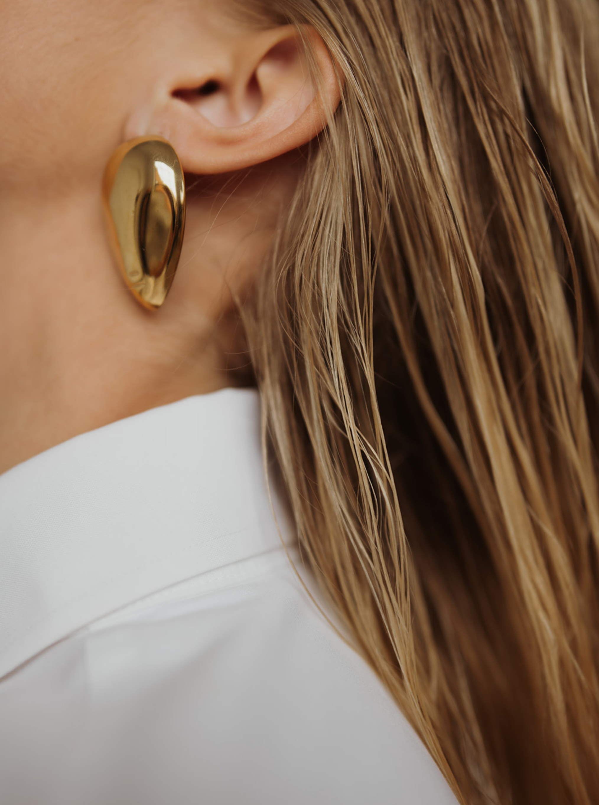 Close-up of a woman's ear with a large, gold heart-shaped earring, part of her white collared shirt, and her long, blonde hair.