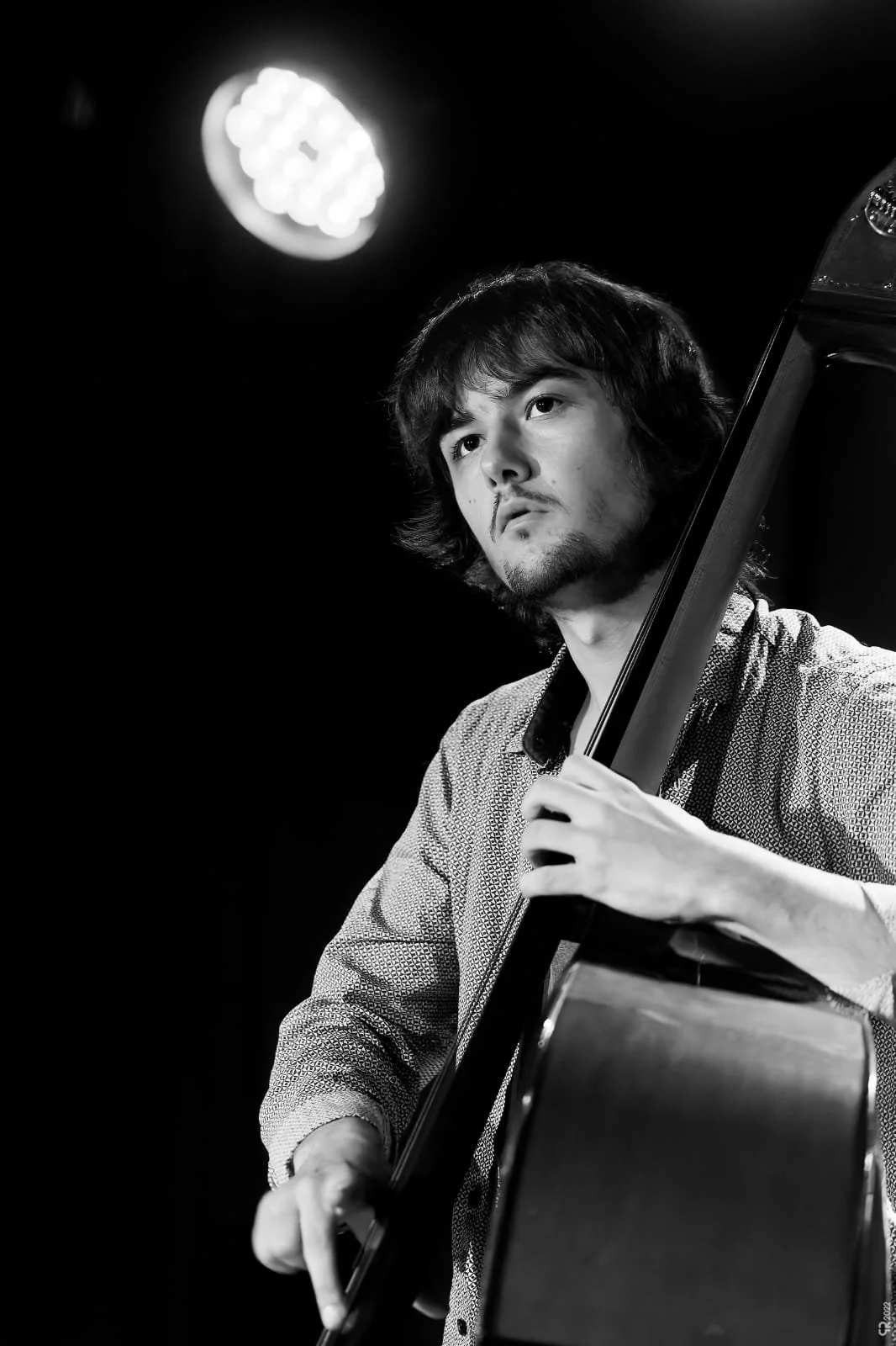 A young man playing an upright bass on stage with a spotlight overhead, black and white photo.