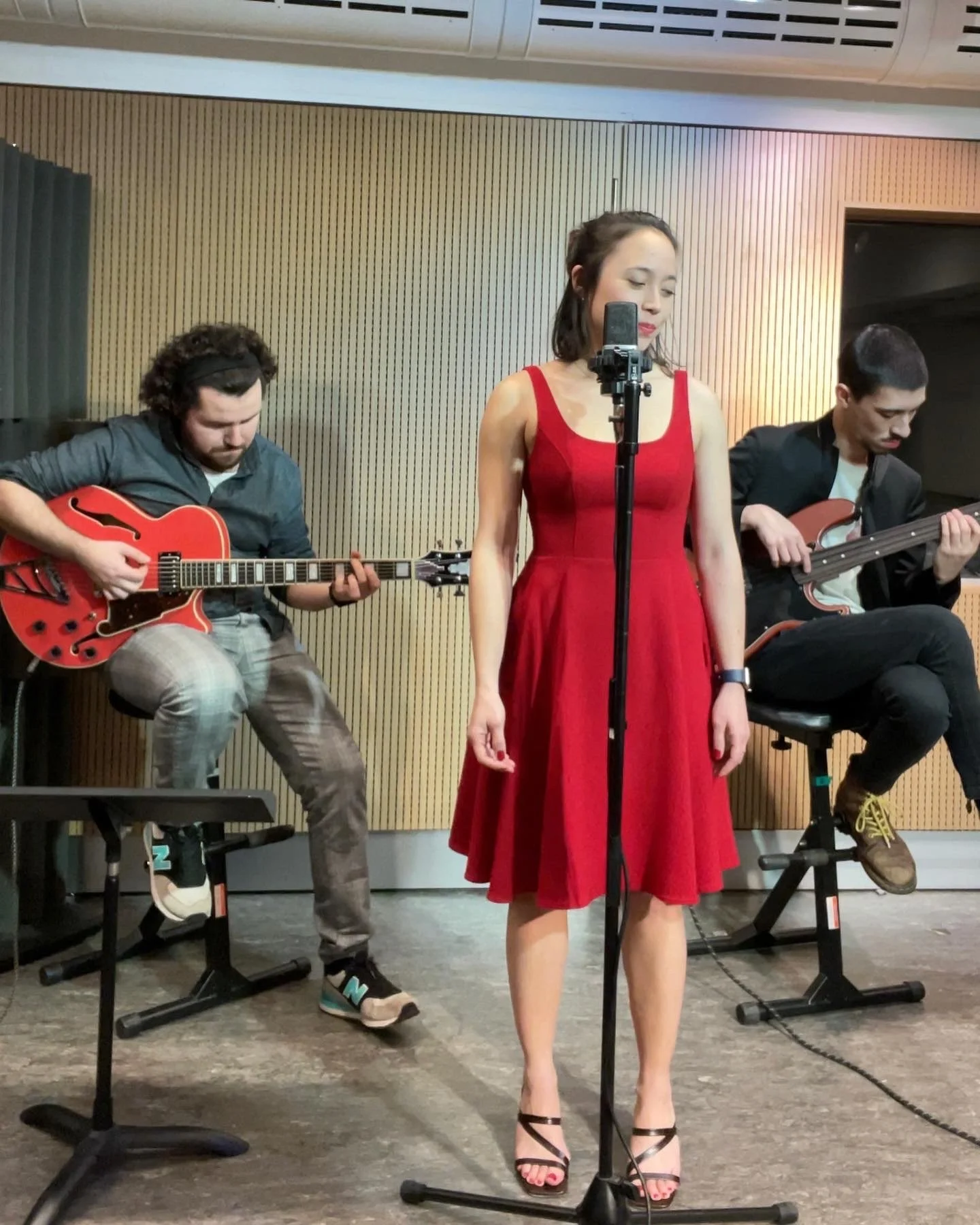 A woman in a red dress singing into a microphone while two men play guitars in the background.