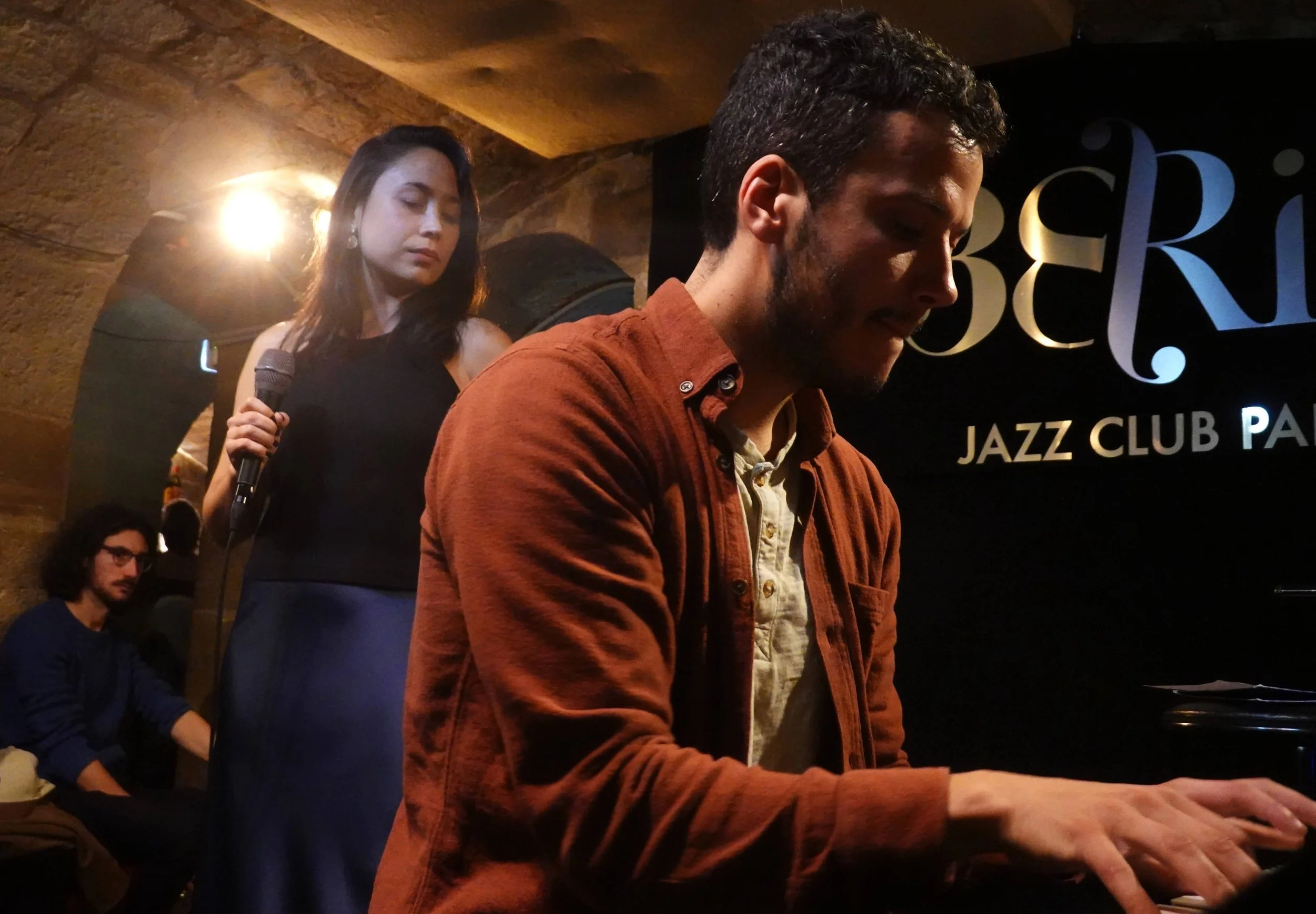 A man playing piano in a jazz club with a woman singing and holding a microphone in the background, and another man sitting in the background.