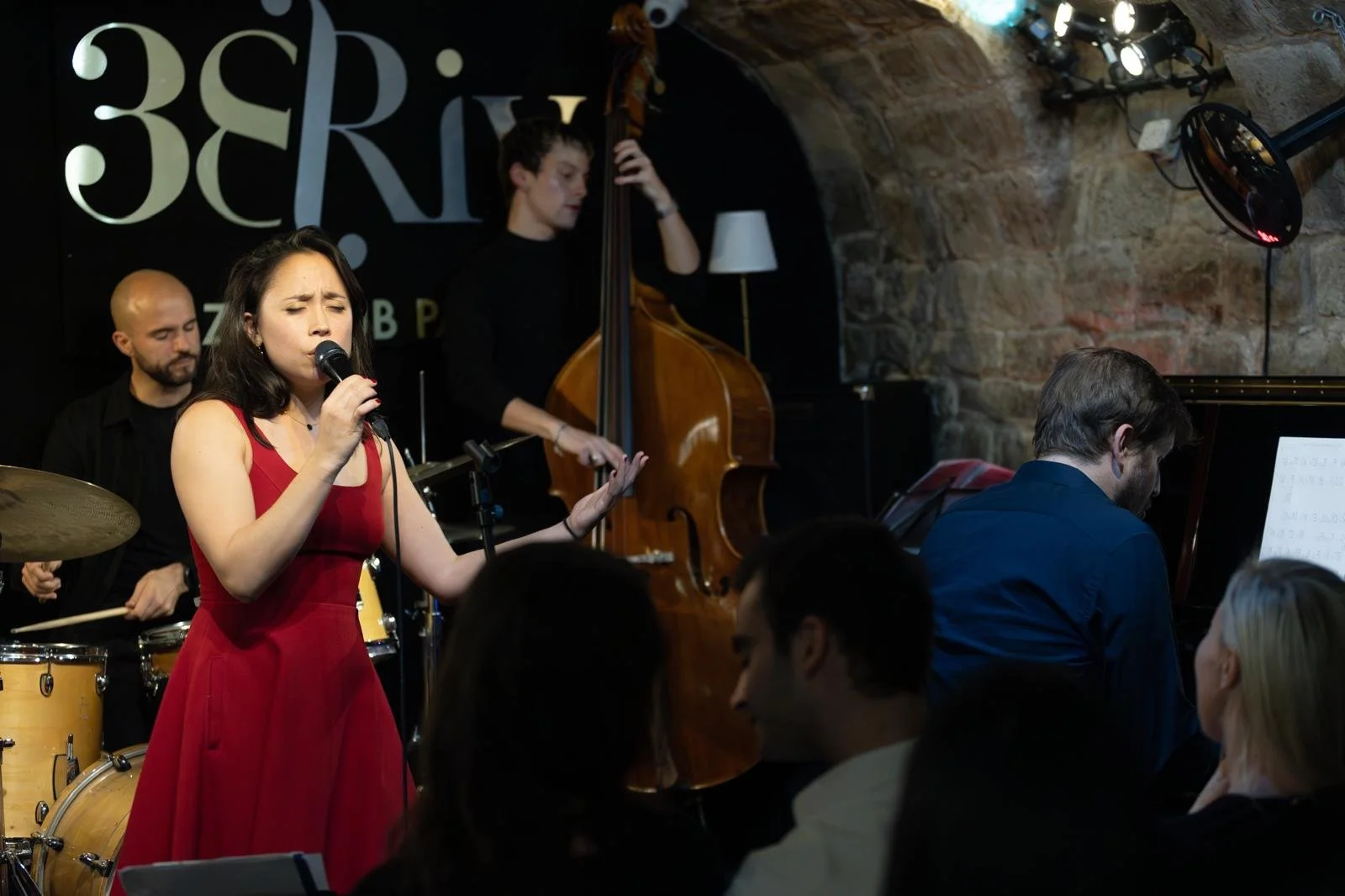 A woman in a red dress is singing into a microphone during a jazz performance at a live music venue. Behind her, a man is playing the drums, a keyboardist is to the right, and a double bassist is in the background. The audience is seated, watching the performance, in a cozy, dimly-lit space with exposed brick walls.