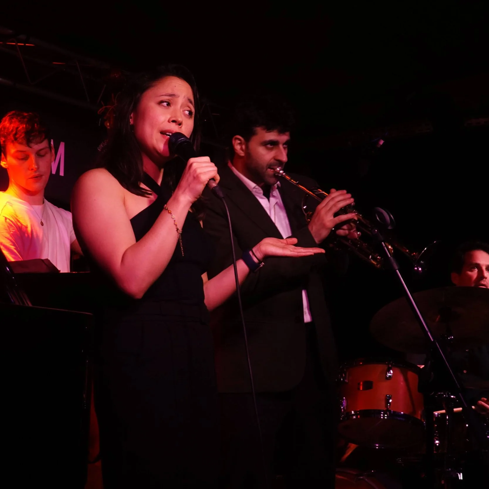 A female singer holding a microphone and a male saxophonist performing on stage, with other band members visible in the background.