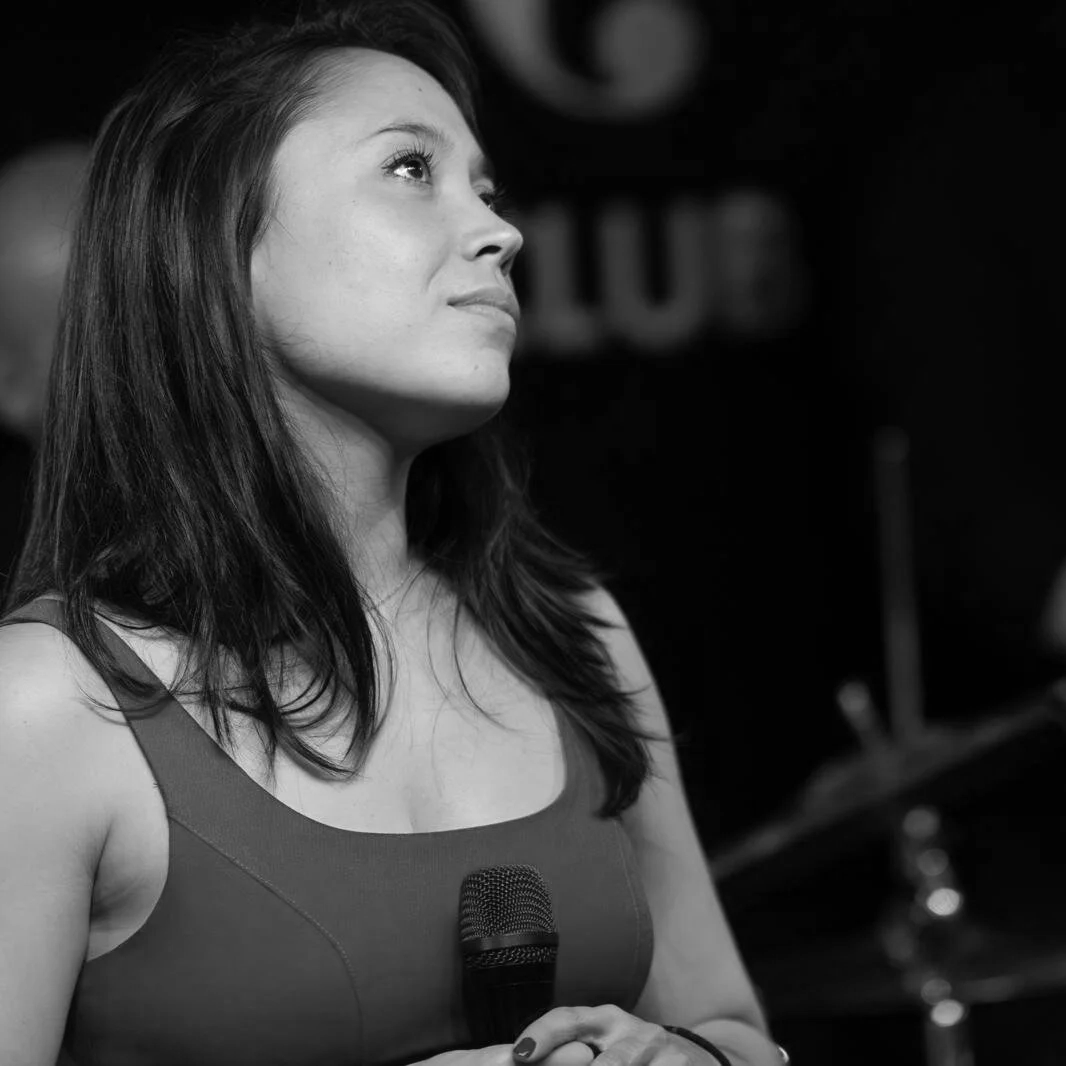 Black and white photo of a woman holding a microphone in her right hand, looking upward and slightly to her left, with long dark hair, wearing a sleeveless top.