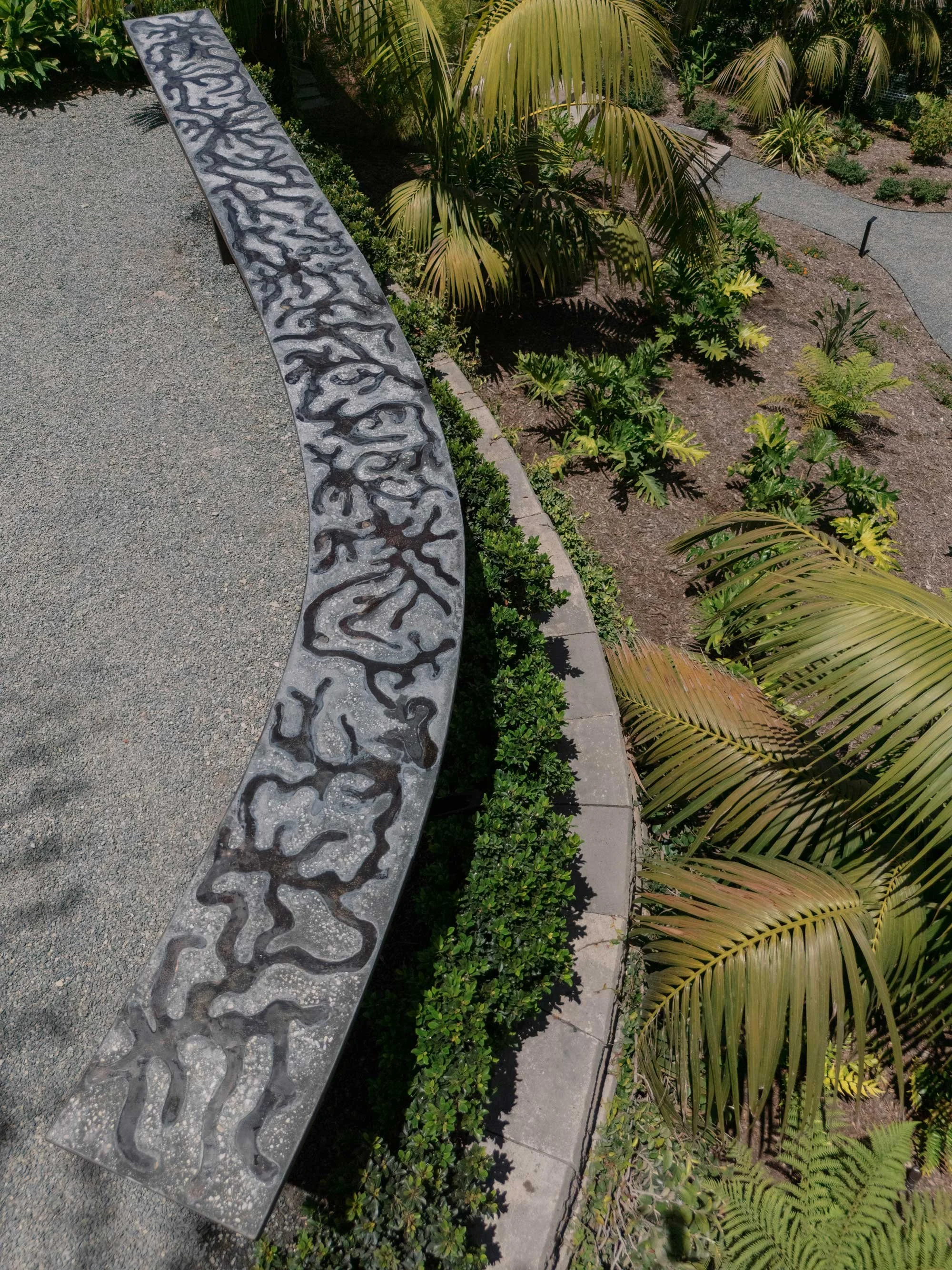 A curved concrete bench with abstract cut-out patterns in a landscaped garden with tropical plants and palm trees.