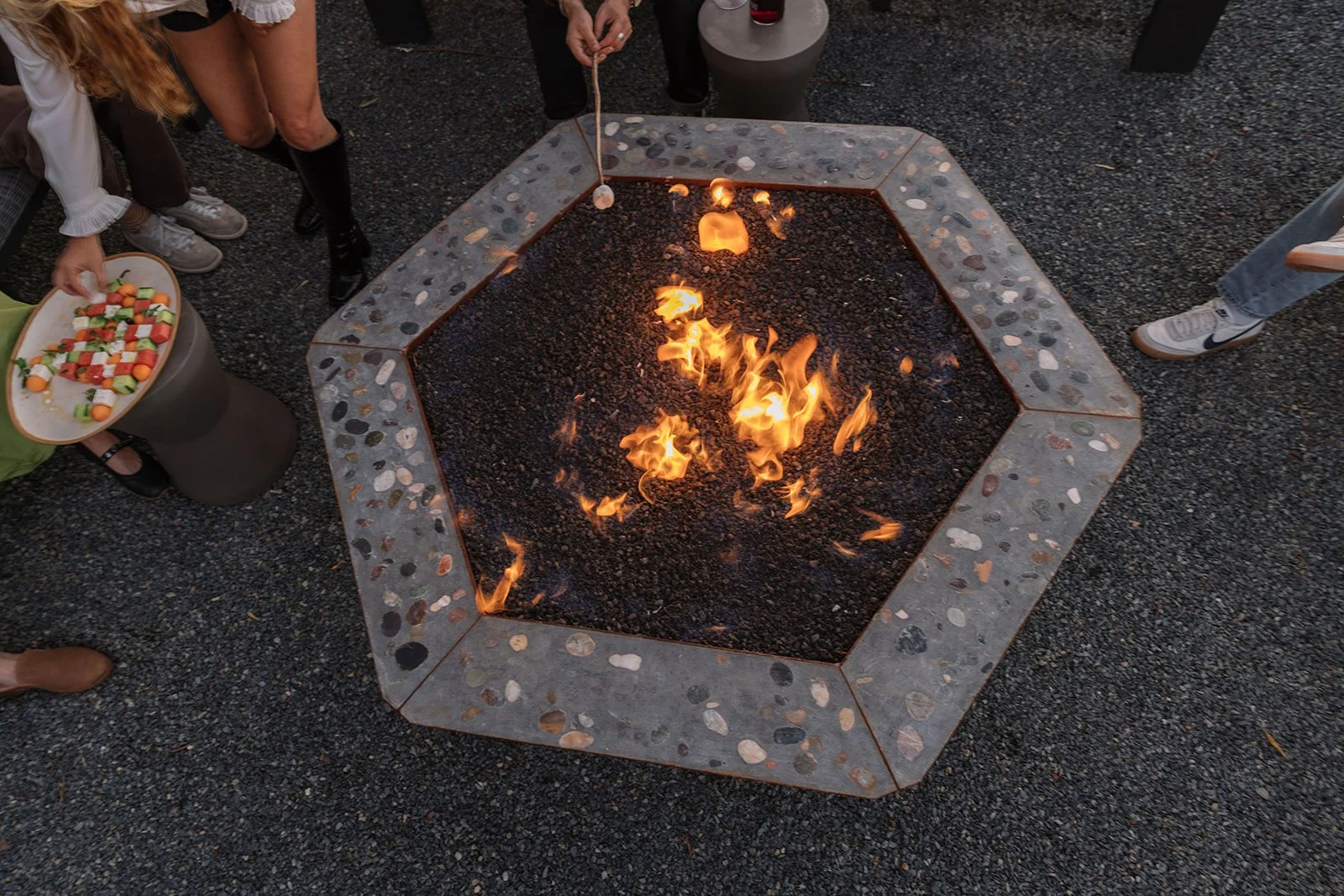 People gathered around a fire pit with a hexagonal concrete border, roasting marshmallows, and a tray of assorted snacks on the side.