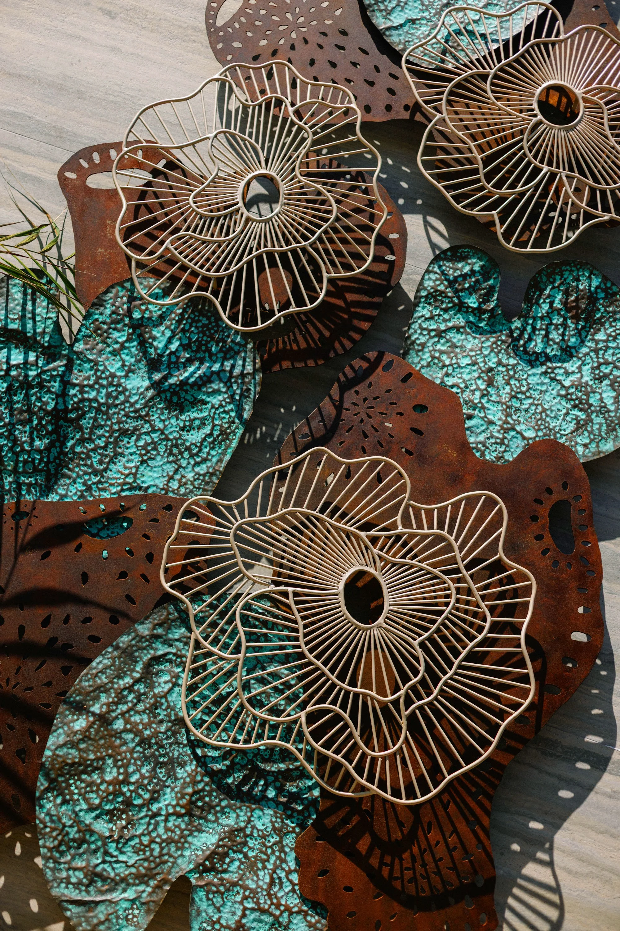 Decorative metal flower-shaped wire baskets and textured blue-green leaves with shadows cast on a light wooden surface.