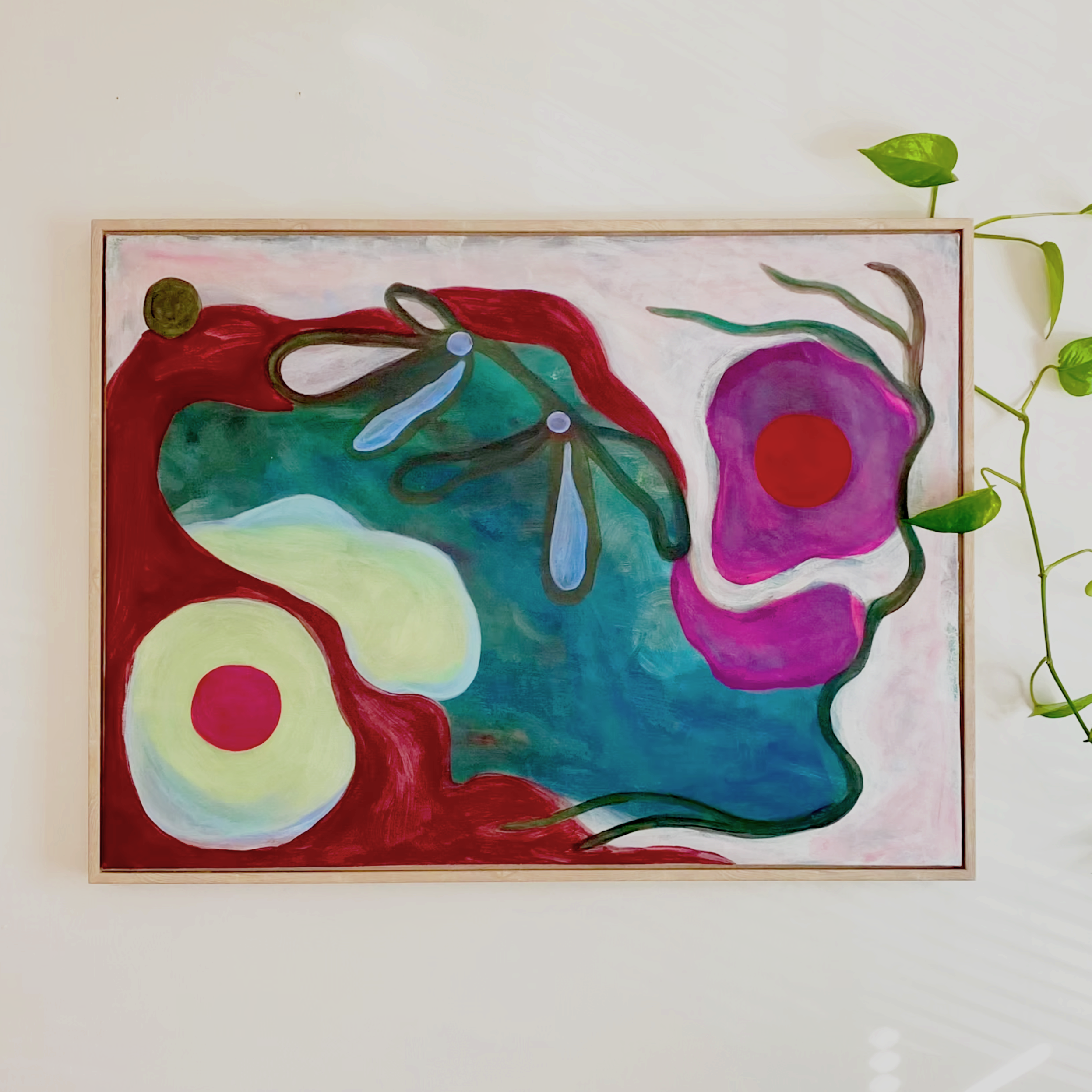 Abstract painting featuring colorful shapes of a face, flowers, and leaves on a white wall, with a leafy plant to the right.