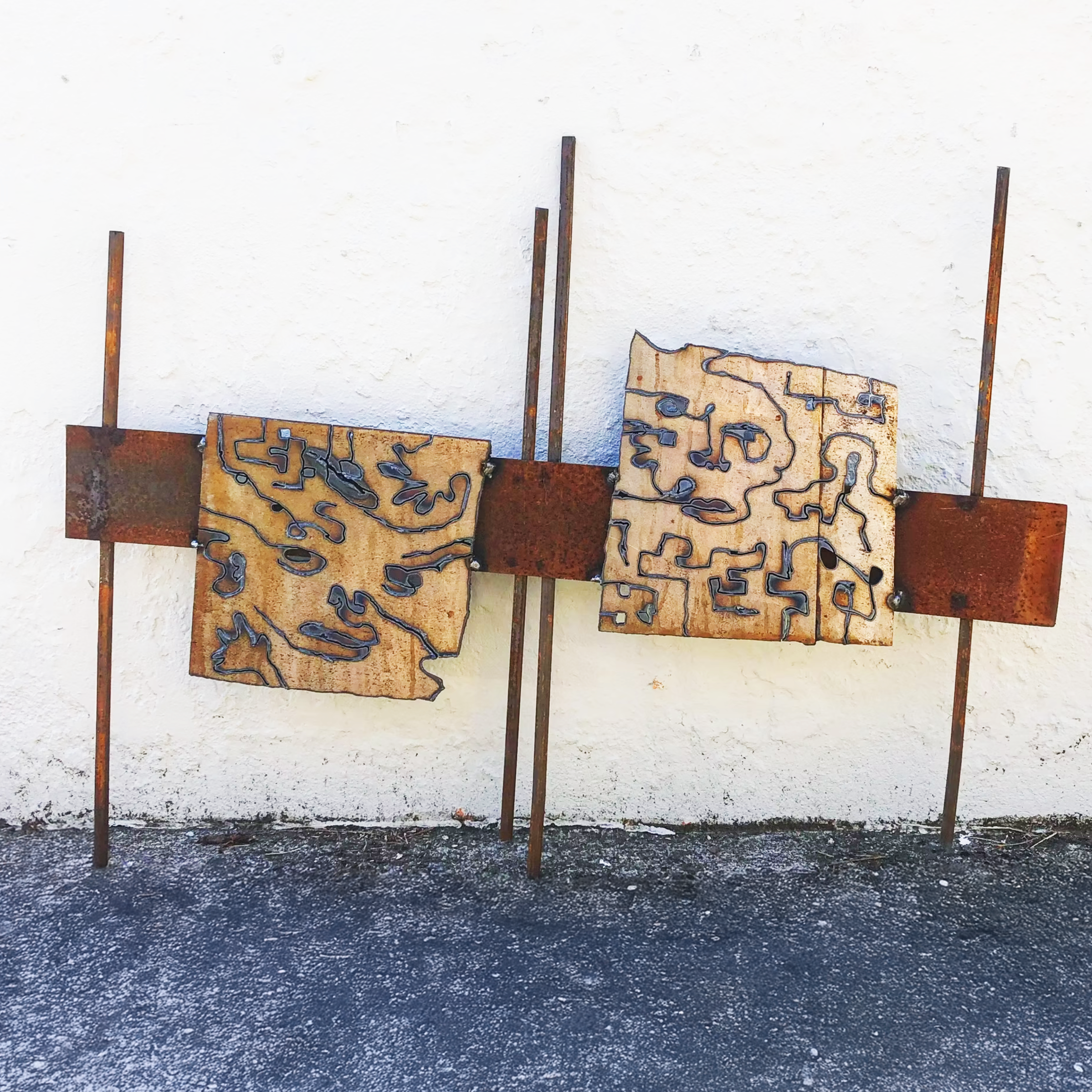 Art installation of two wooden panels with black maze-like patterns, mounted on a rusted metal structure against a white wall.