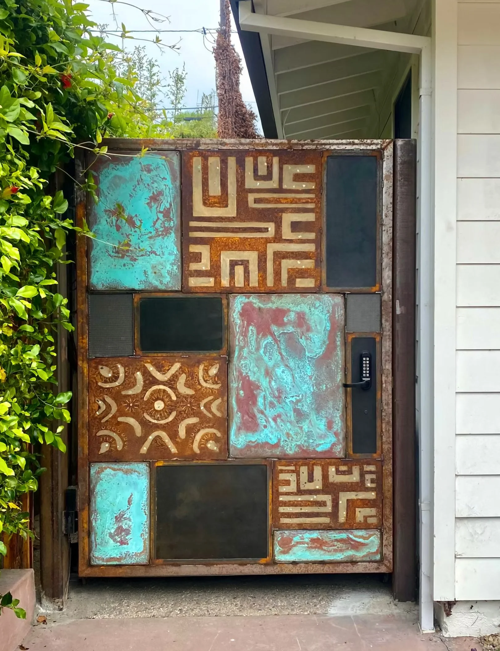 Decorative gate with a mix of rust, turquoise, black, and patterned metal panels, and a keypad lock, positioned between a green leafy bush and a white house exterior.