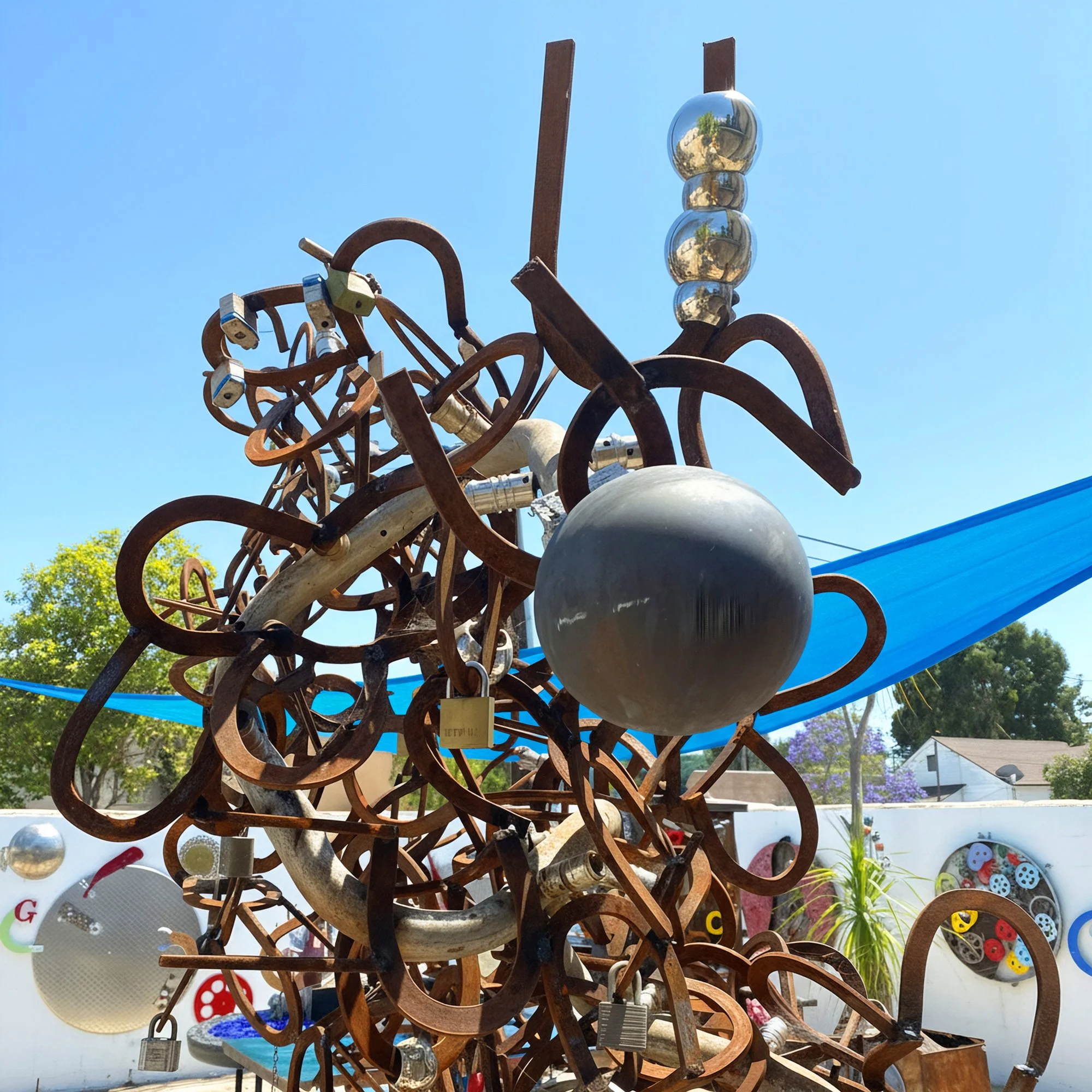 An abstract outdoor sculpture made of intertwined metal and wooden pieces with reflective spheres and padlocks, under a clear blue sky.