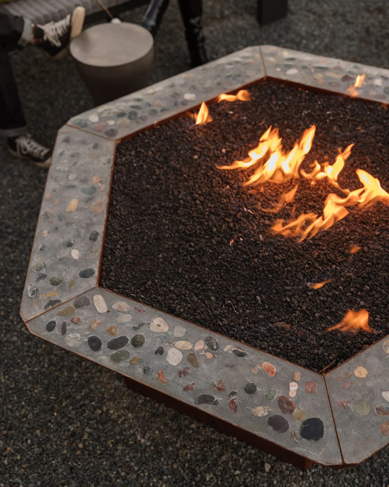 Square fire pit with a stone border, filled with dark lava rocks and small flames, outdoors at dusk.