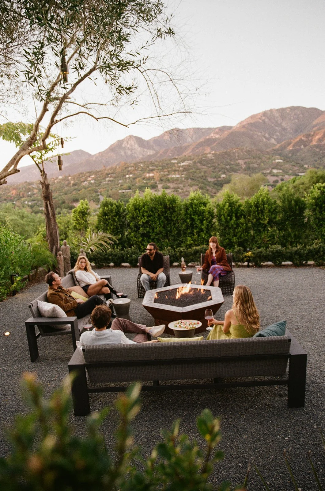 Group of seven people sitting around a fire pit outdoors, with mountains and green trees in the background, enjoying drinks and conversation at dusk.