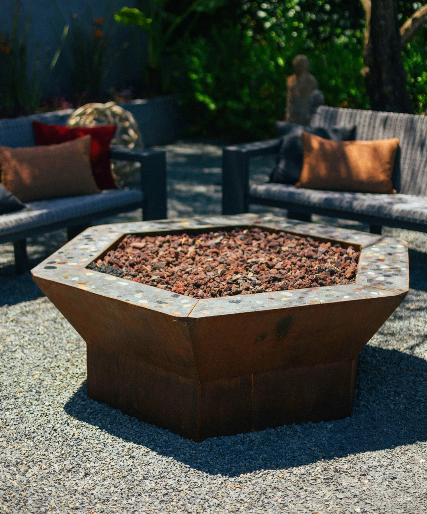Outdoor patio with a modern fire pit filled with small rocks, surrounded by wicker sofas with colorful cushions, lush green plants, and a small stone sculpture in the background.