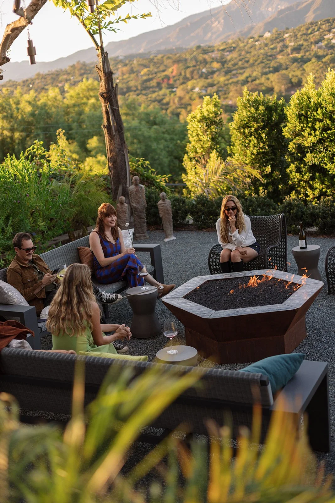 A group of five friends enjoying a fire pit gathering in an outdoor patio surrounded by greenery and mountains in the background during late afternoon or early evening.