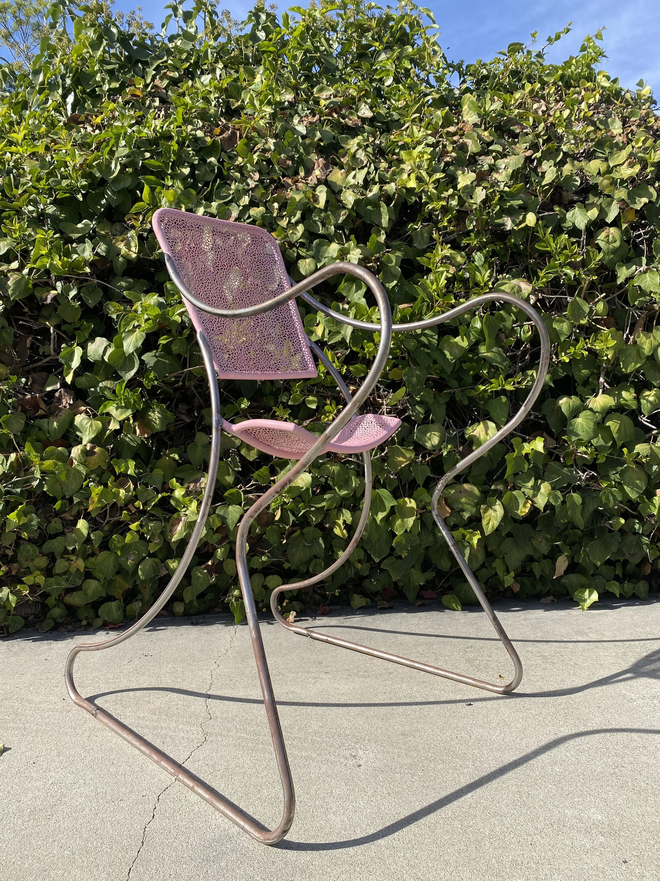 A pink modern chair with a mesh seat and backrest, with a curved metal frame, placed outdoors on a concrete surface with a green leafy bush in the background and a blue sky.