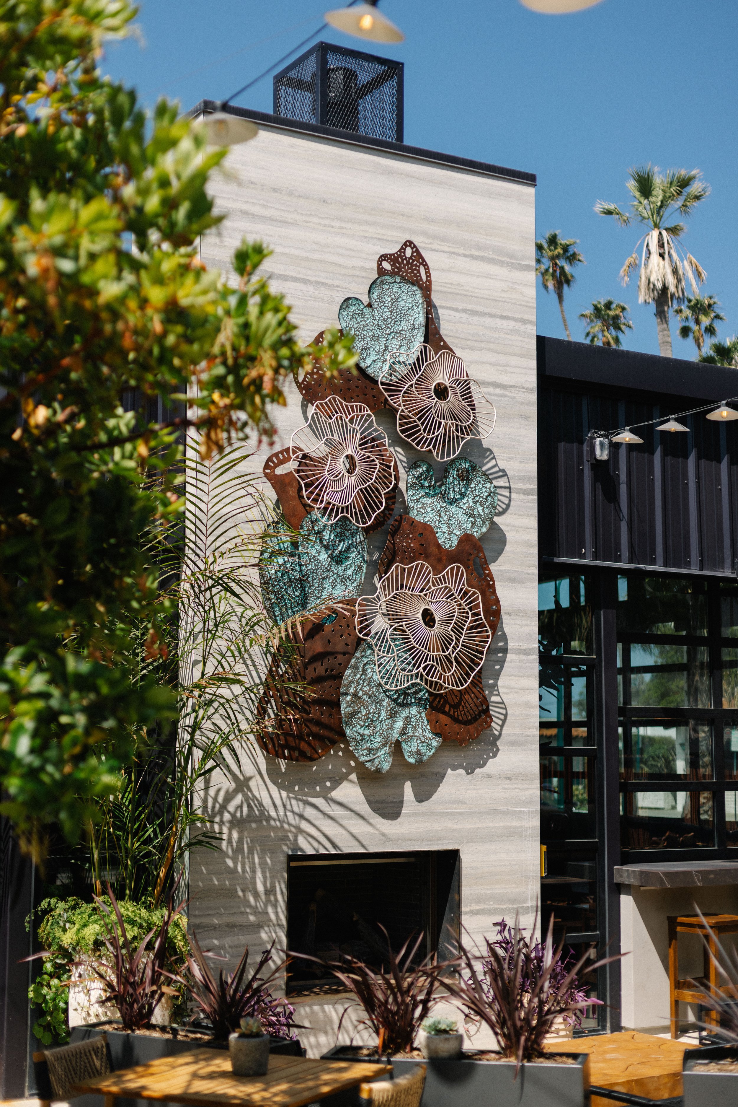 Exterior wall with artistic metal wall sculpture featuring flowers and abstract shapes, set on a modern building with large windows and surrounding plants.