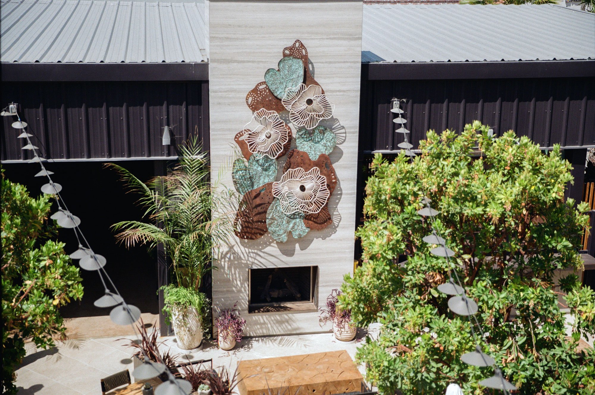 Outdoor courtyard with potted plants, decorative metal wall art of leaves and flowers on a white wall, a small fireplace, surrounded by trees and string lights.