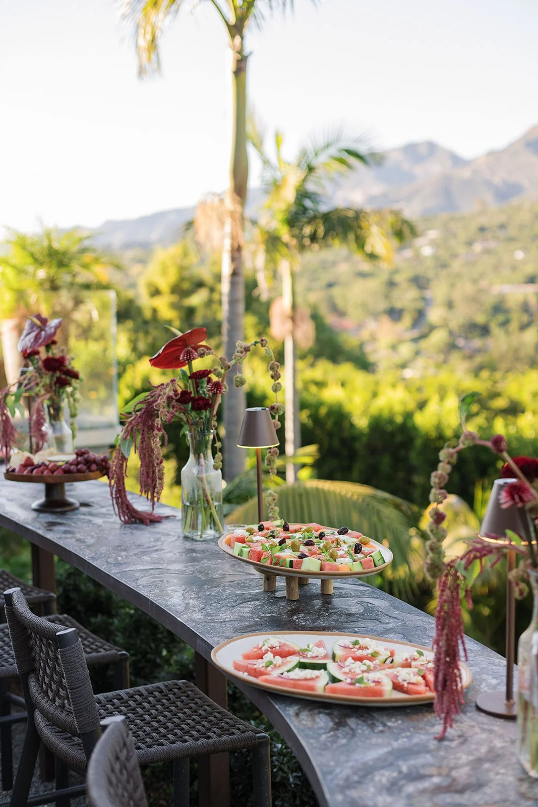 Outdoor buffet table with fruit platters and floral arrangements, overlooking a lush green landscape with mountains and palm trees in the background.