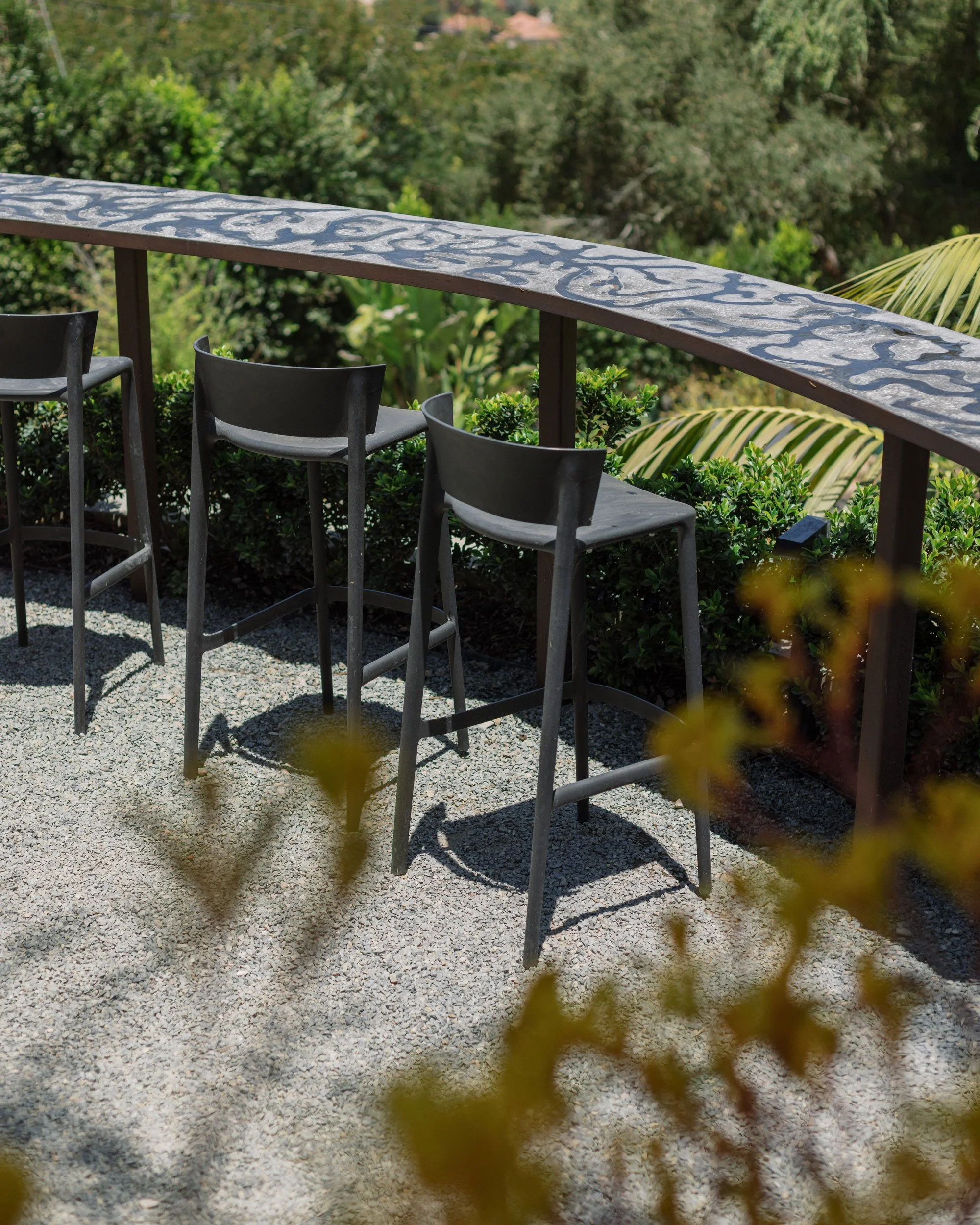 Outdoor balcony with black metal bar stools and a decorative railing, surrounded by green bushes and trees.