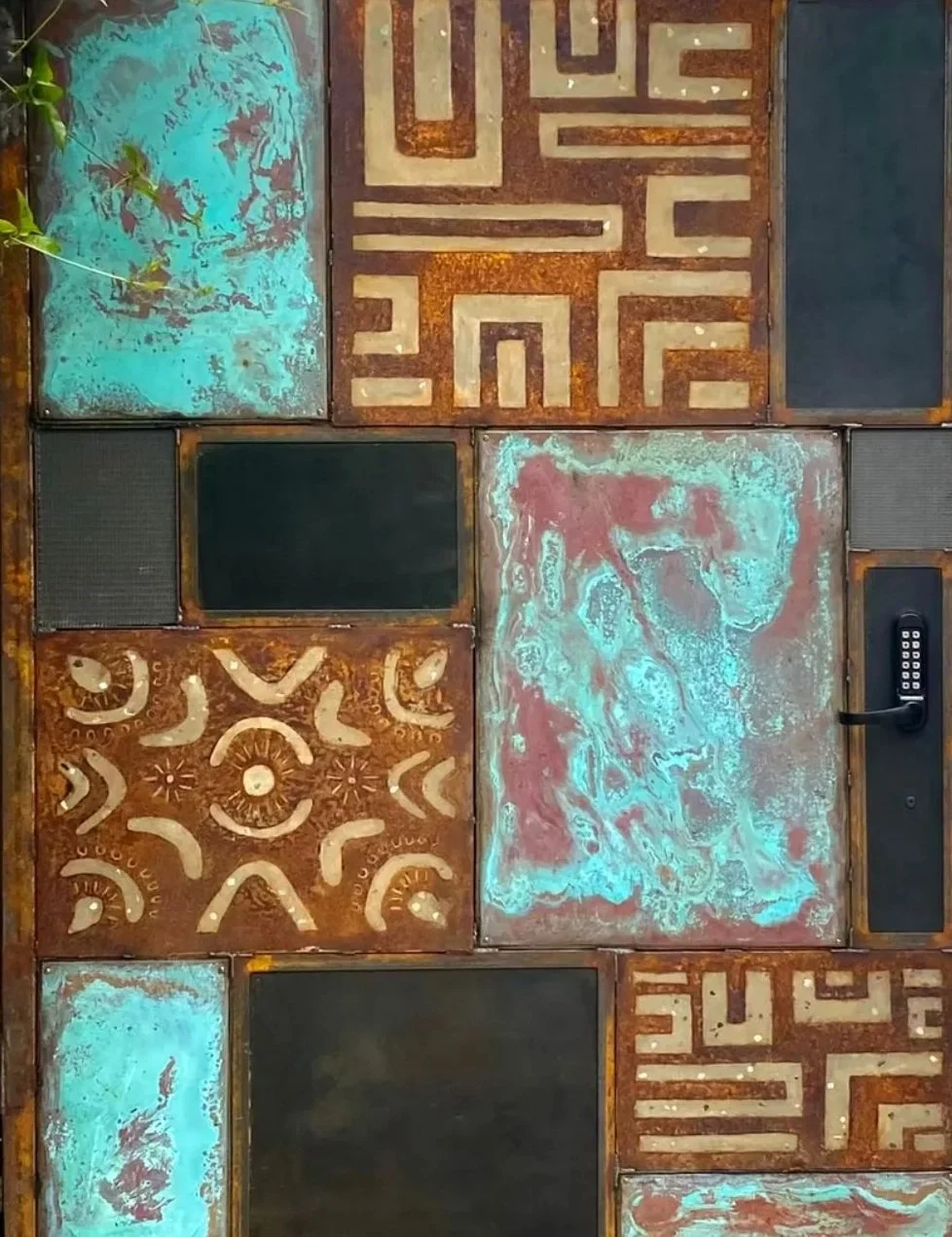 A wall decorated with various square panels featuring different geometric and abstract patterns, some with rusted metal textures and others with painted designs, including a lion's face and maze-like patterns. One panel includes a black keypad lock.