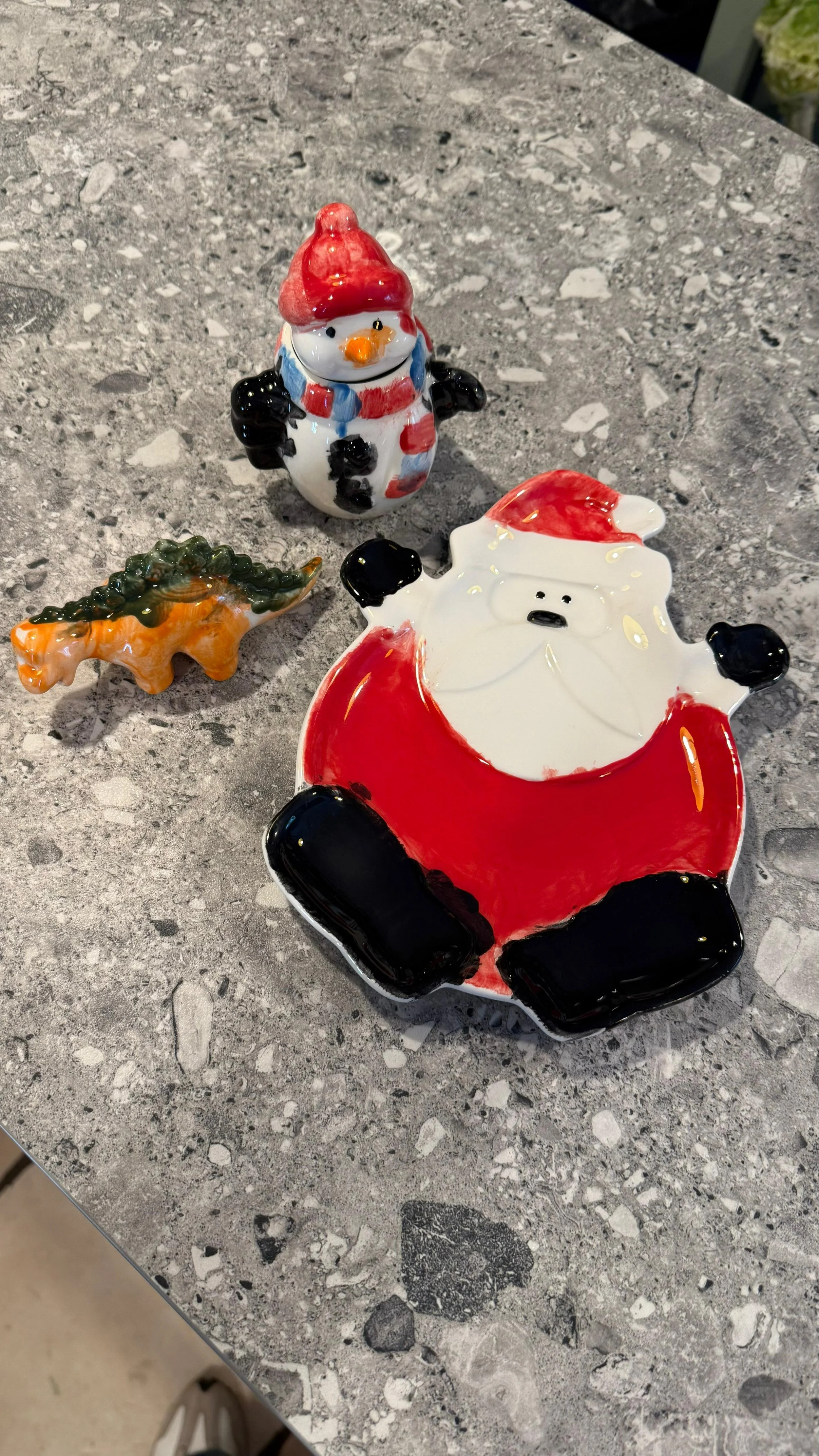 Colorful ceramic holiday decorations including a snowman, Santa Claus, and a small Christmas tree on a speckled gray countertop.