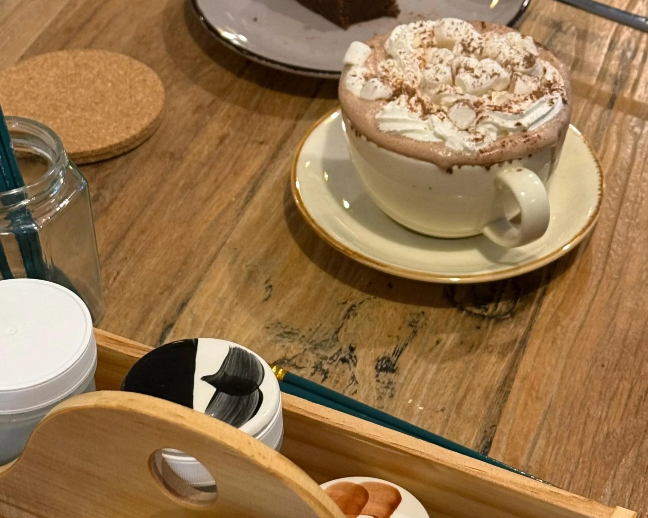 A cup of hot chocolate topped with whipped cream and marshmallows, served on a saucer on a wooden table.