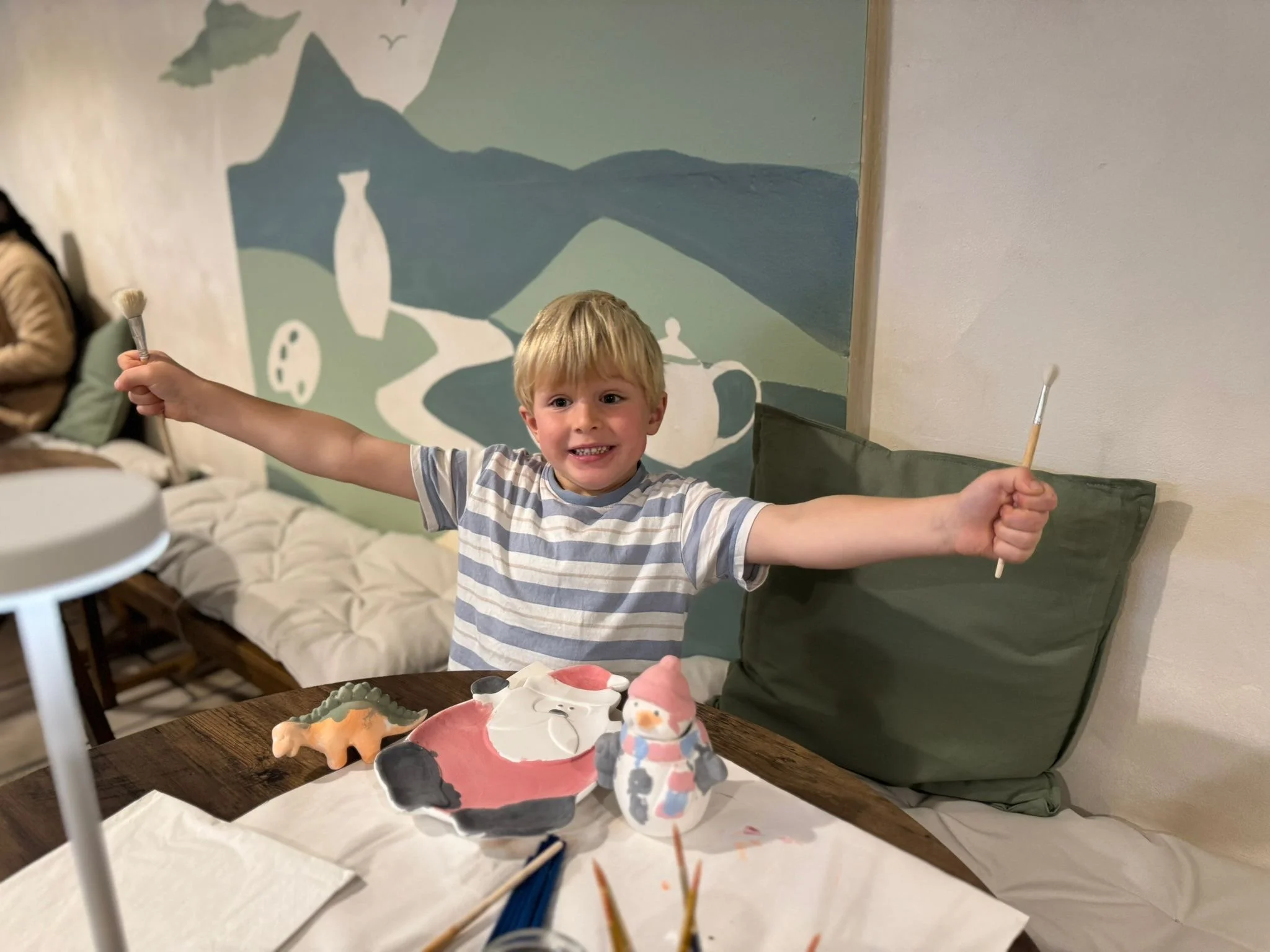 A young boy with blond hair smiling happily, holding a paintbrush in each hand with arms outstretched. He is sitting at a table with Christmas-themed painted ceramic decorations, including a Santa Claus and a snowman. Behind him, there is a colorful wall mural with abstract mountain and landscape shapes.