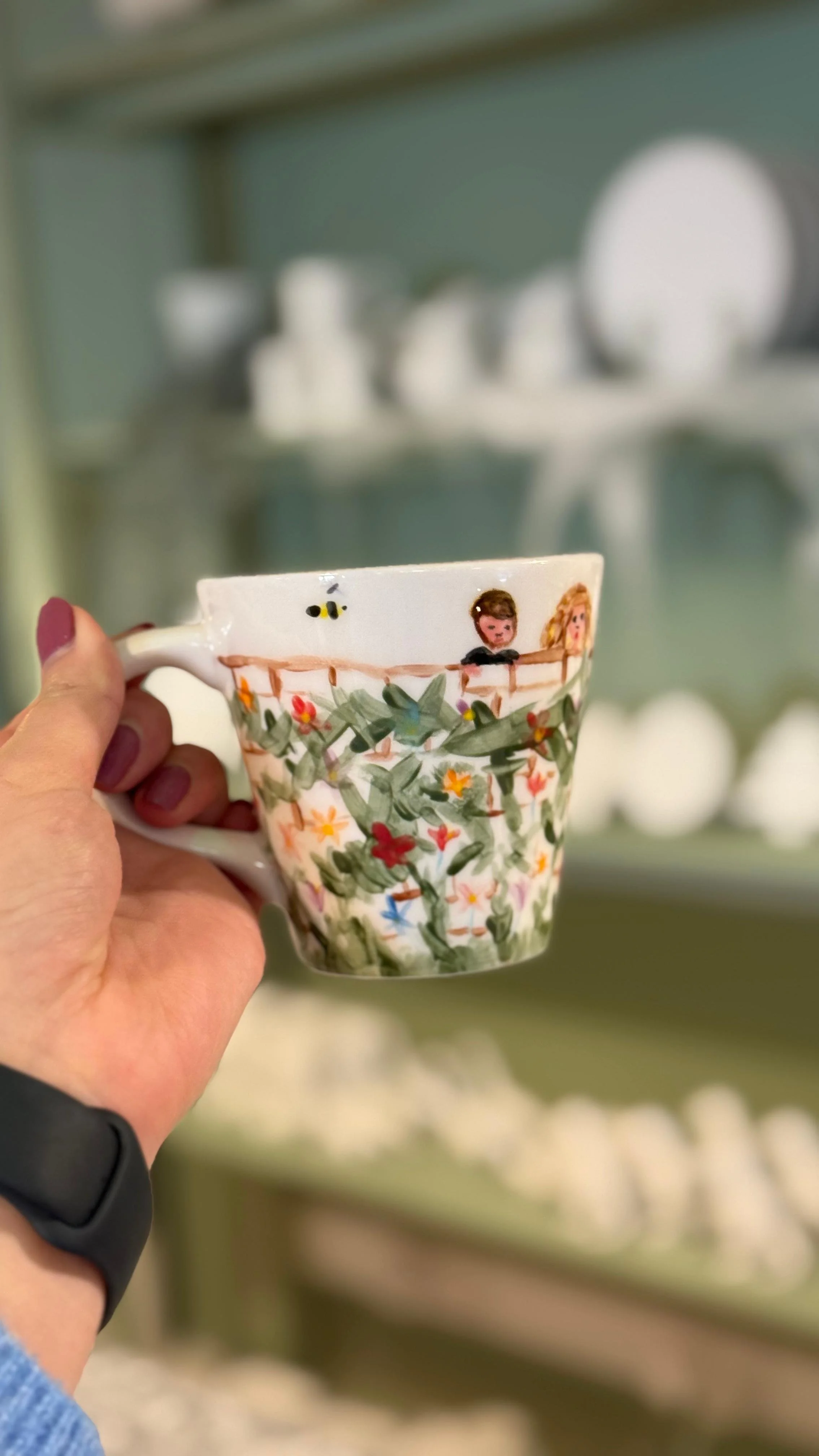 A person holding a decorated ceramic mug with a floral pattern and cartoon characters, with a blurred background of additional ceramic items.
