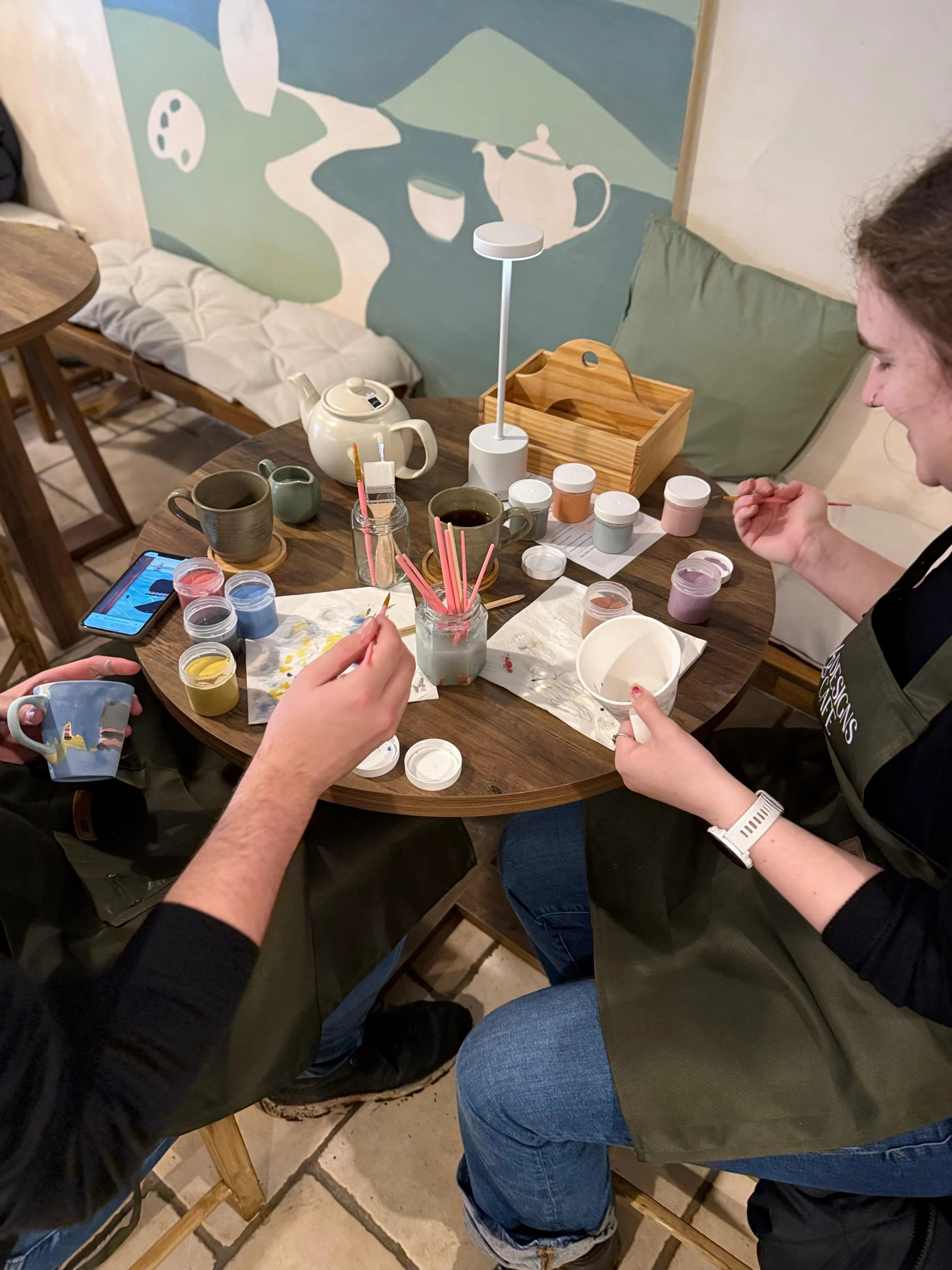 Two people sitting at a round wooden table, painting ceramics with small jars of paint, brushes, and cups of water in a cozy room with a mural on the wall and a wooden bench with cushions.