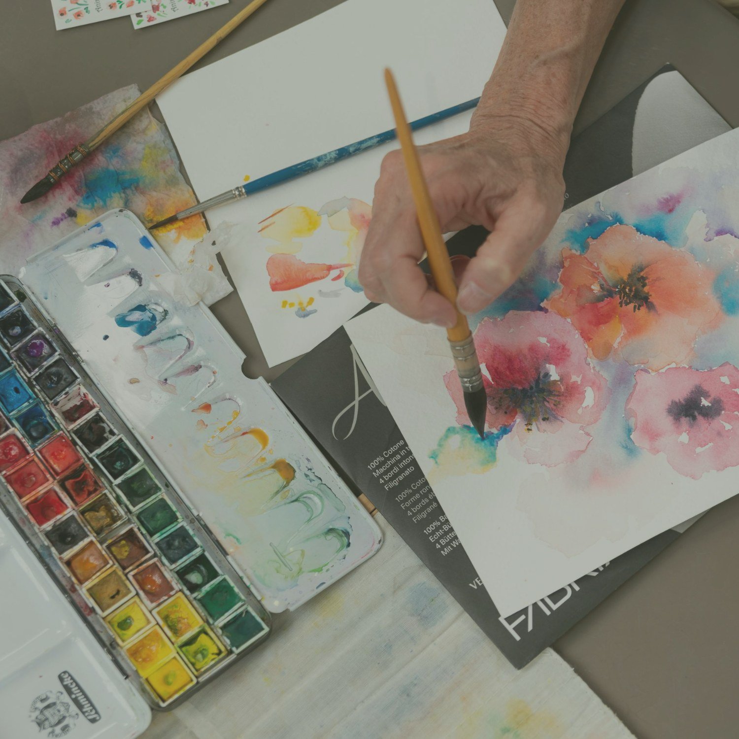 Hands painting pink watercolour flowers next to a watercolour palette at Designs Café, Dartington