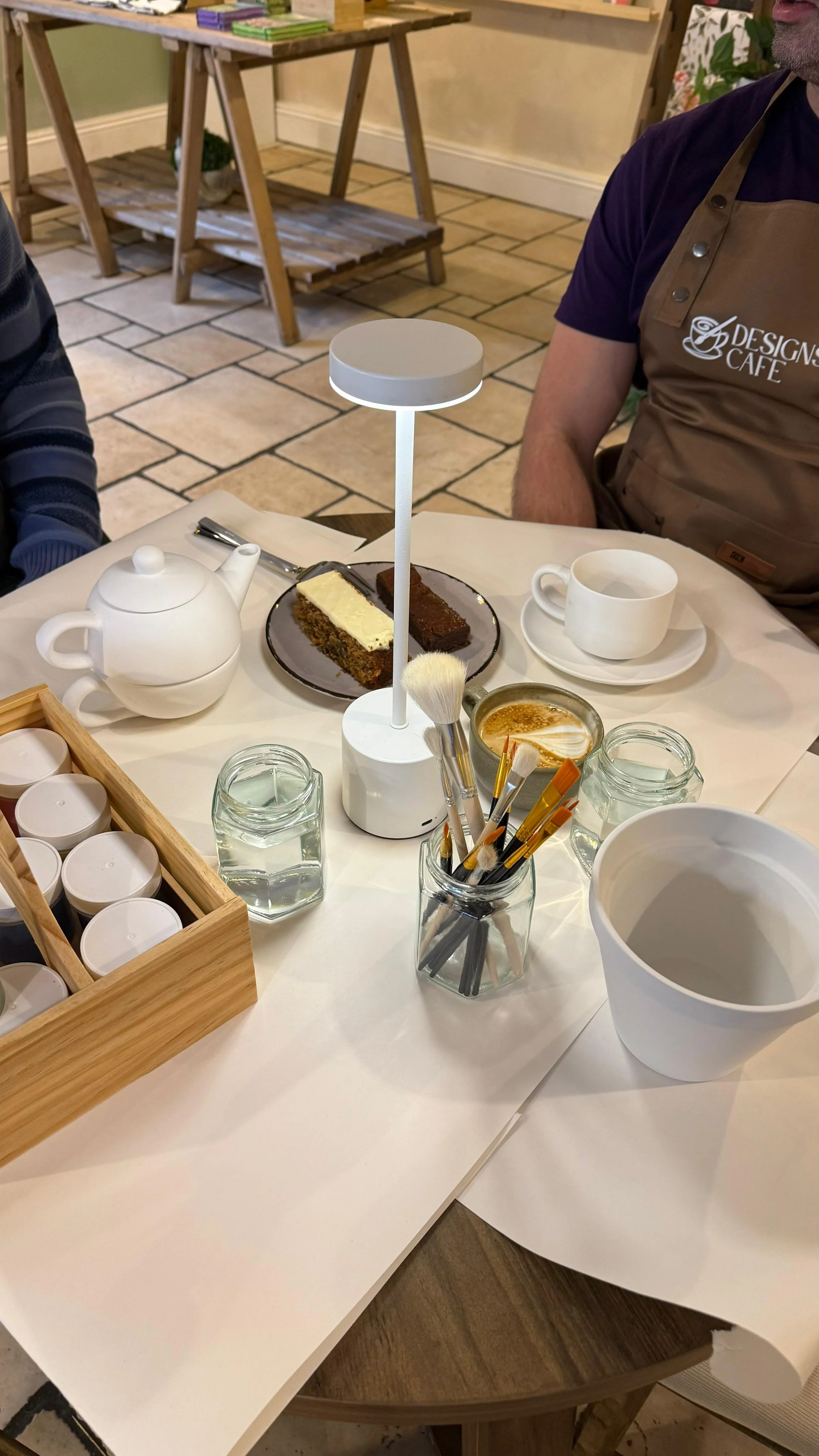 A table in a cafe with a teapot, two cakes, a white coffee cup, a glass of water, some brushes, jars, and a display light.