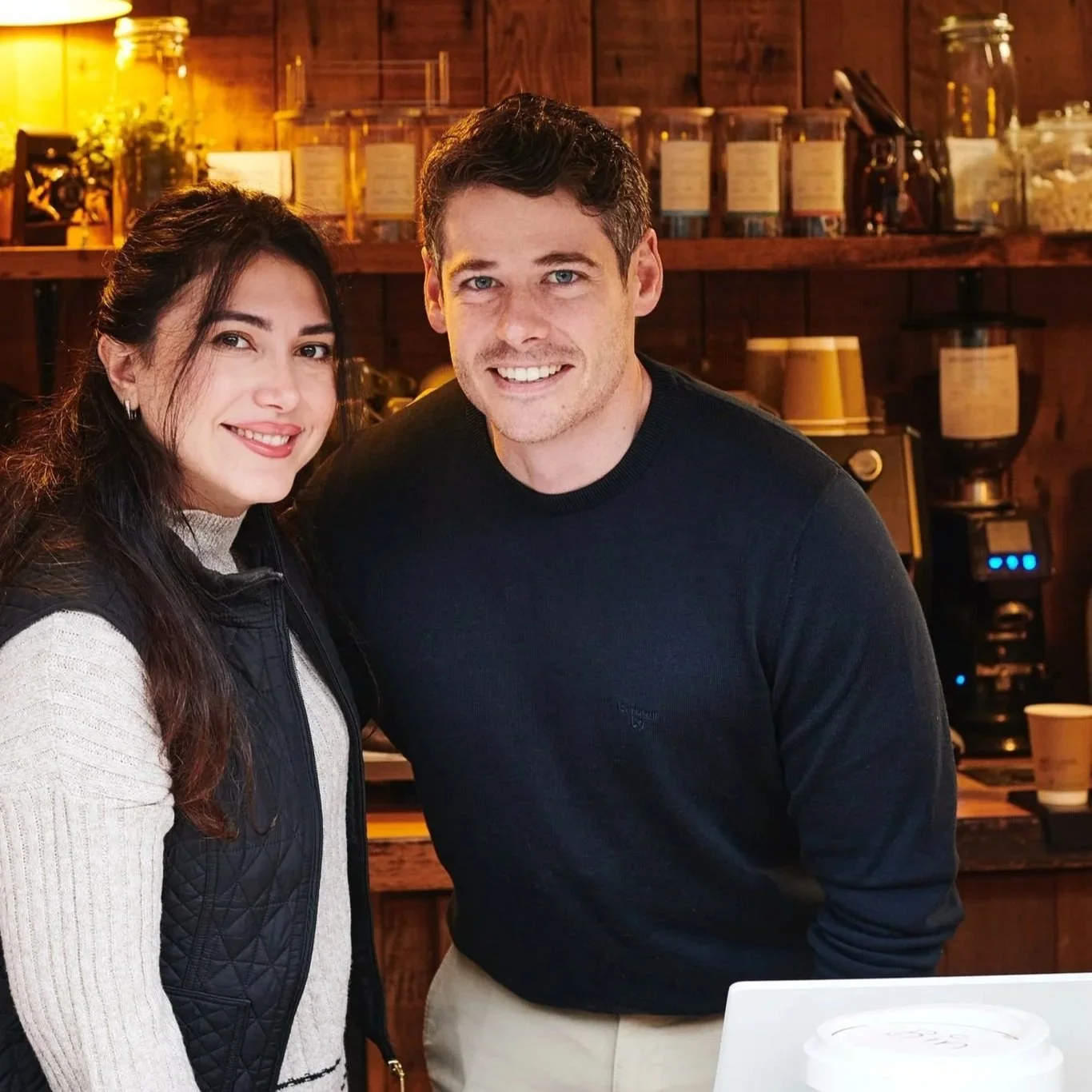 A smiling man and woman standing together inside a cozy, warmly-lit coffee shop or cafe.