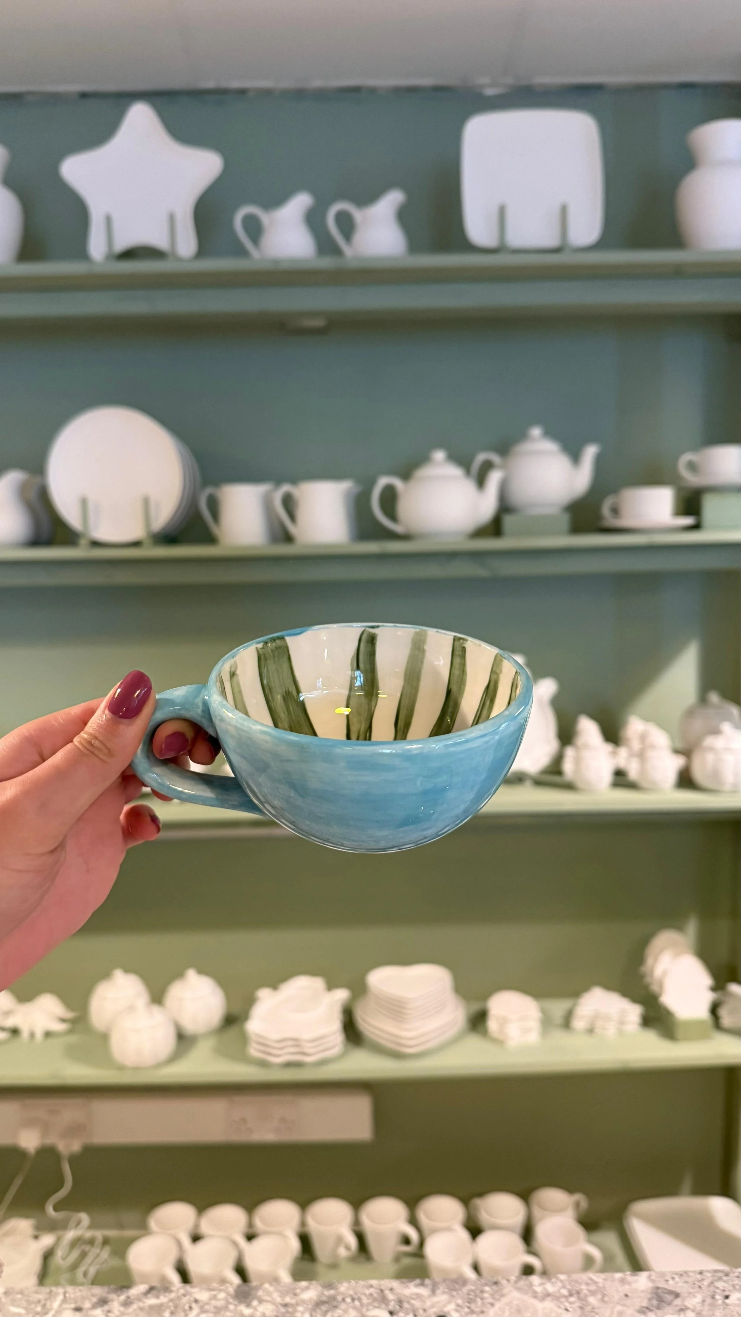 Person holding a blue ceramic bowl with green and white striped interior in front of shelves filled with white ceramic dinnerware set.
