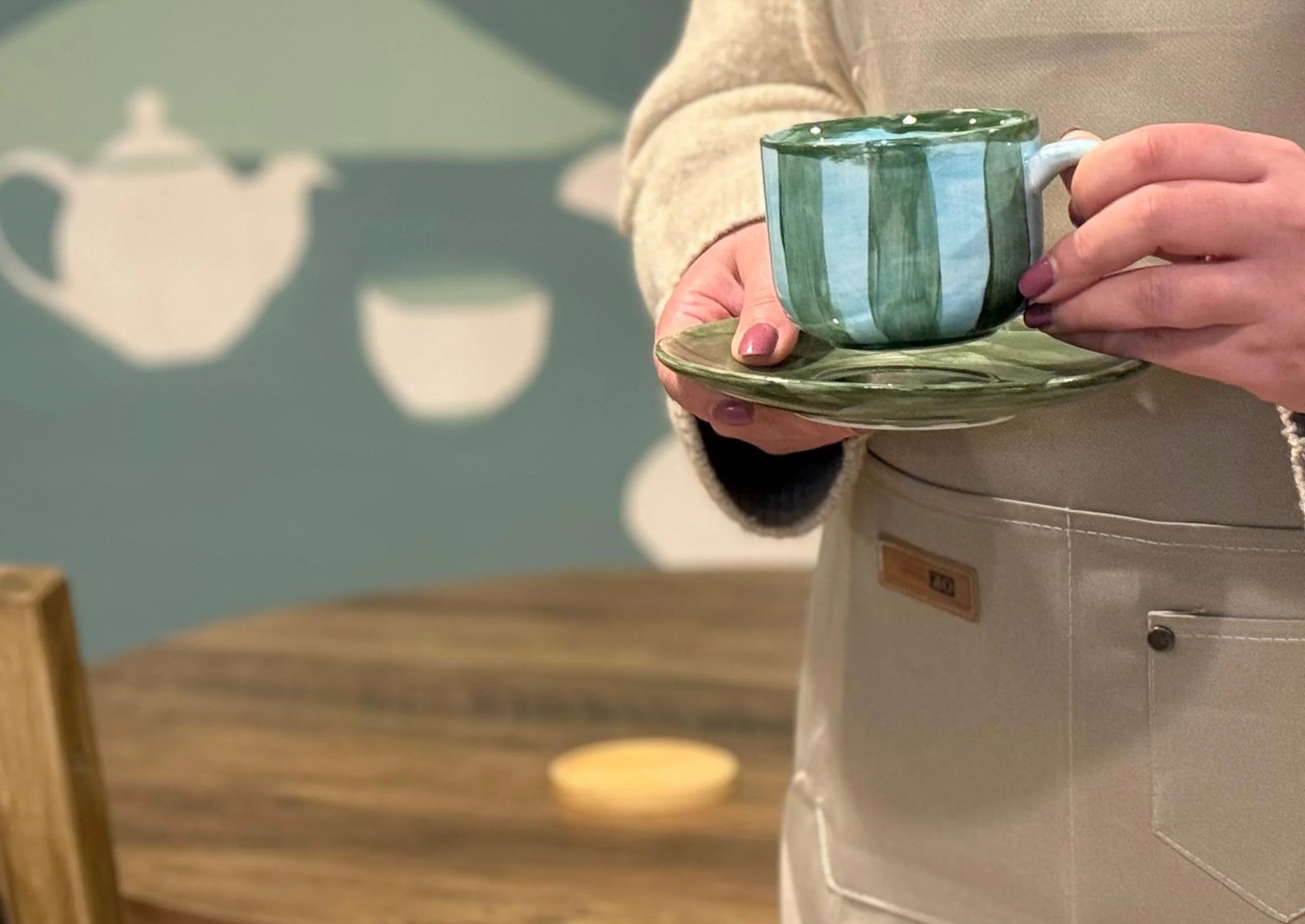 Person holding a ceramic mug and saucer in a kitchen or cafe setting.