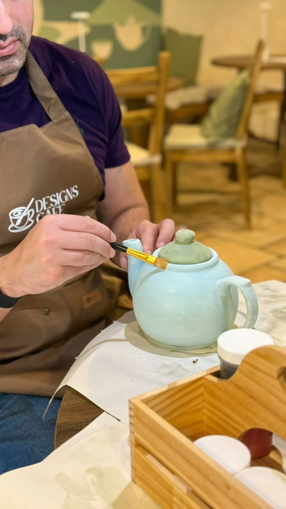 A painter in a Designs Café apron brushing yellow detail onto a pale green teapot during a pottery painting session
