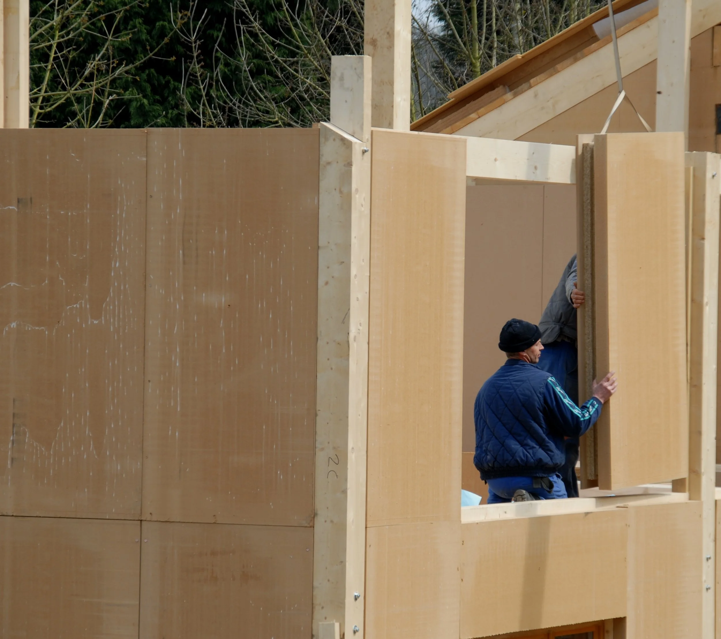 Deux hommes construisent une maison en bois, une personne en veste grise et bonnet, l'autre en veste bleue, en train de fixer une grande planche en bois.