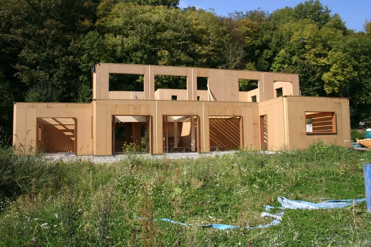 Une maison en Sanomur et sa structure poteaux poutres, avec un cadre non fini, située dans un environnement naturel avec de l'herbe et des arbres en arrière-plan.