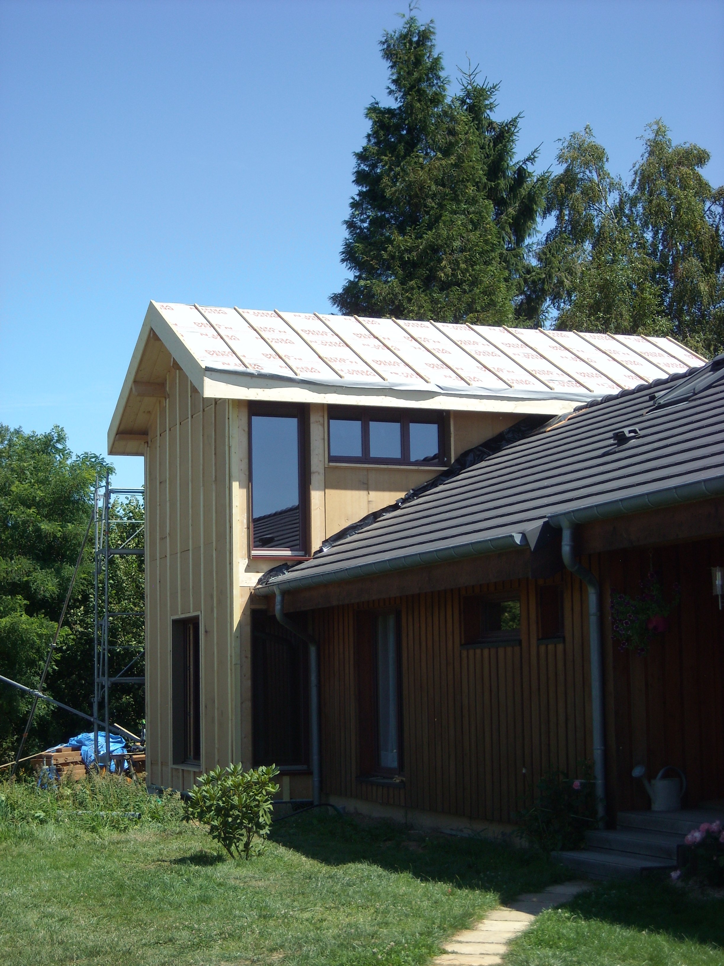 Une maison en cours de construction avec un toit en partie installé, des fenêtres modernes, une façade en bois clair, un jardin avec un chemin en pierre, et des arbres en arrière-plan.