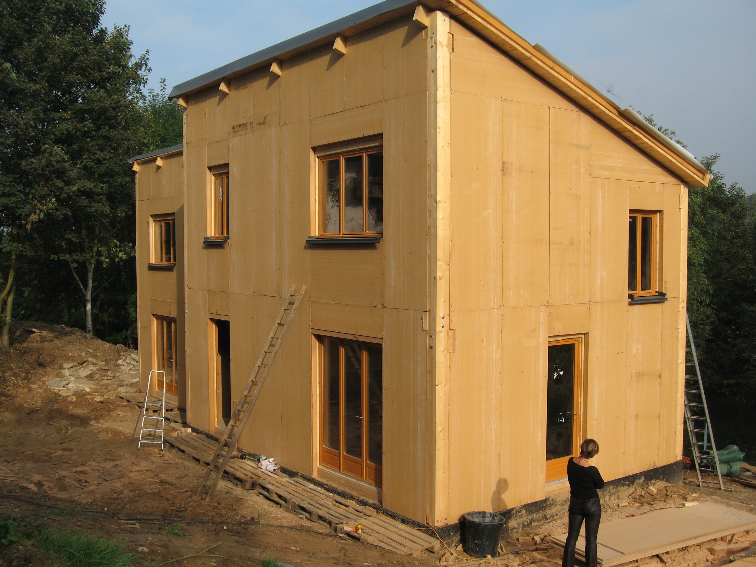 Maison en construction en bois avec plusieurs fenêtres, un personnen regarde le chantier, des escabeaux et des matériaux de construction