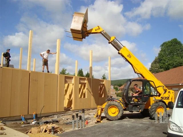 Une grue jaune soulève un grand container en bois sur un chantier de construction, avec plusieurs ouvriers à l'étage en train de travailler ou d'observer. Le ciel est partiellement nuageux et un bâtiment avec un toit rouge est visible en arrière-plan
