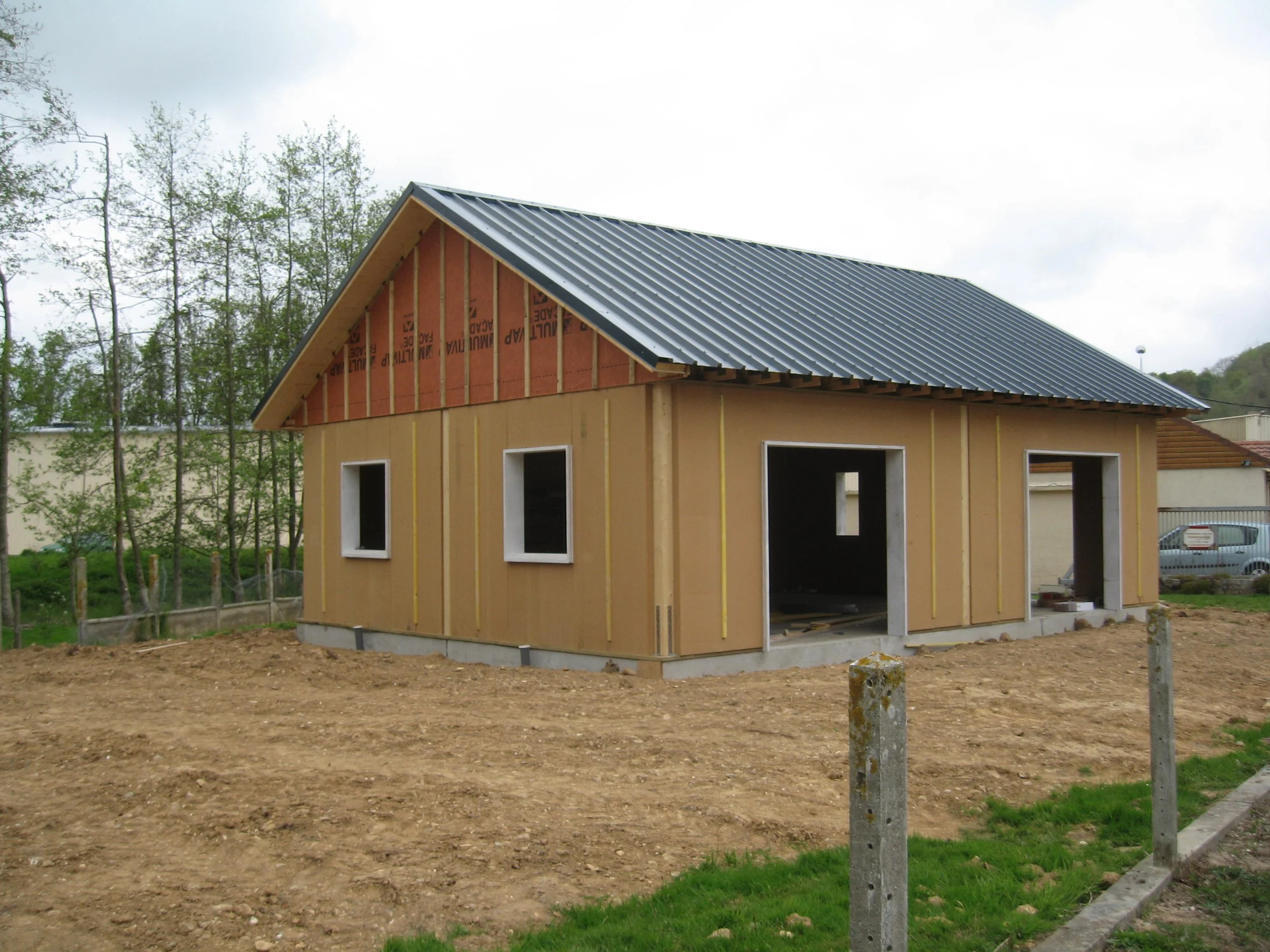 Maison en construction avec murs en bois et toiture en métal, sans fenêtres ni porte installées, sur un terrain en terre, avec des arbres et d'autres bâtiments en arrière-plan.
