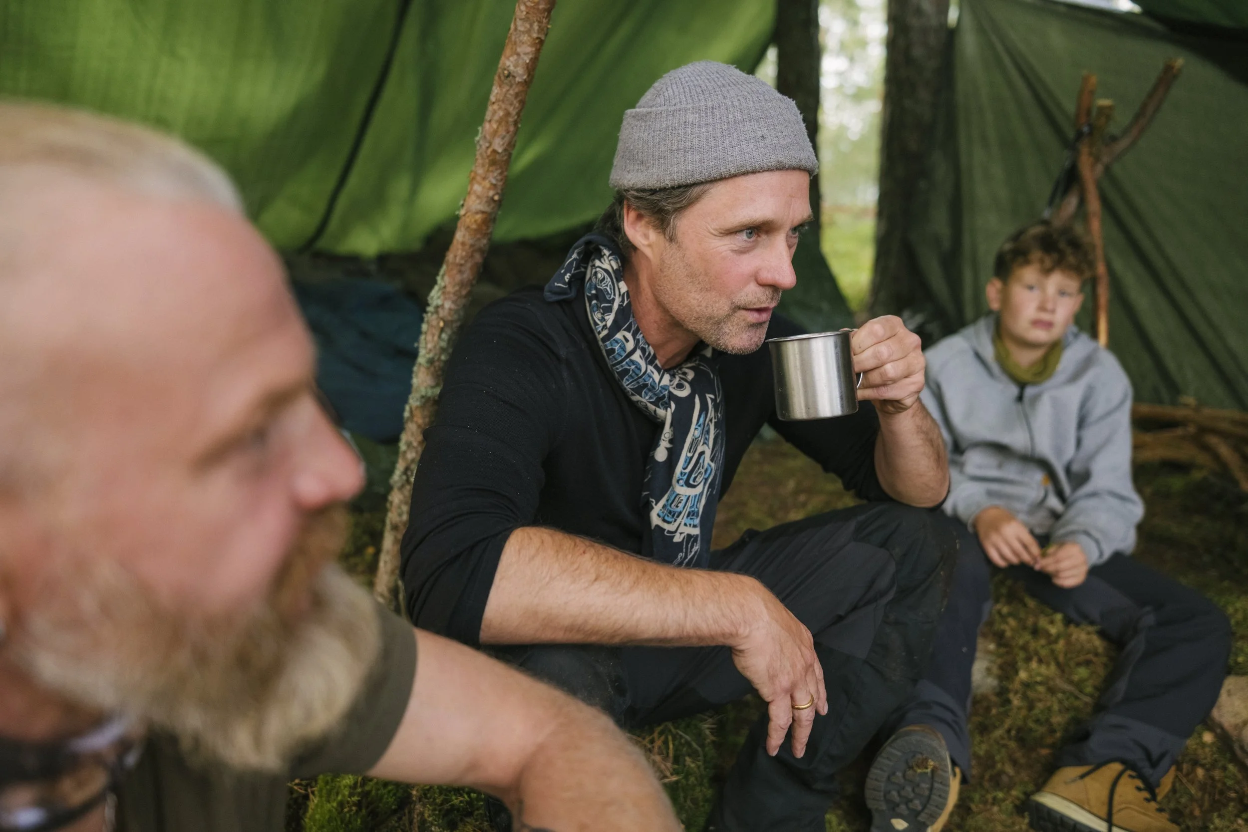 Drei Menschen sitzen in einem Wald, ein Mann trinkt aus einer Metalltasse, während zwei Jungen daneben sitzen.