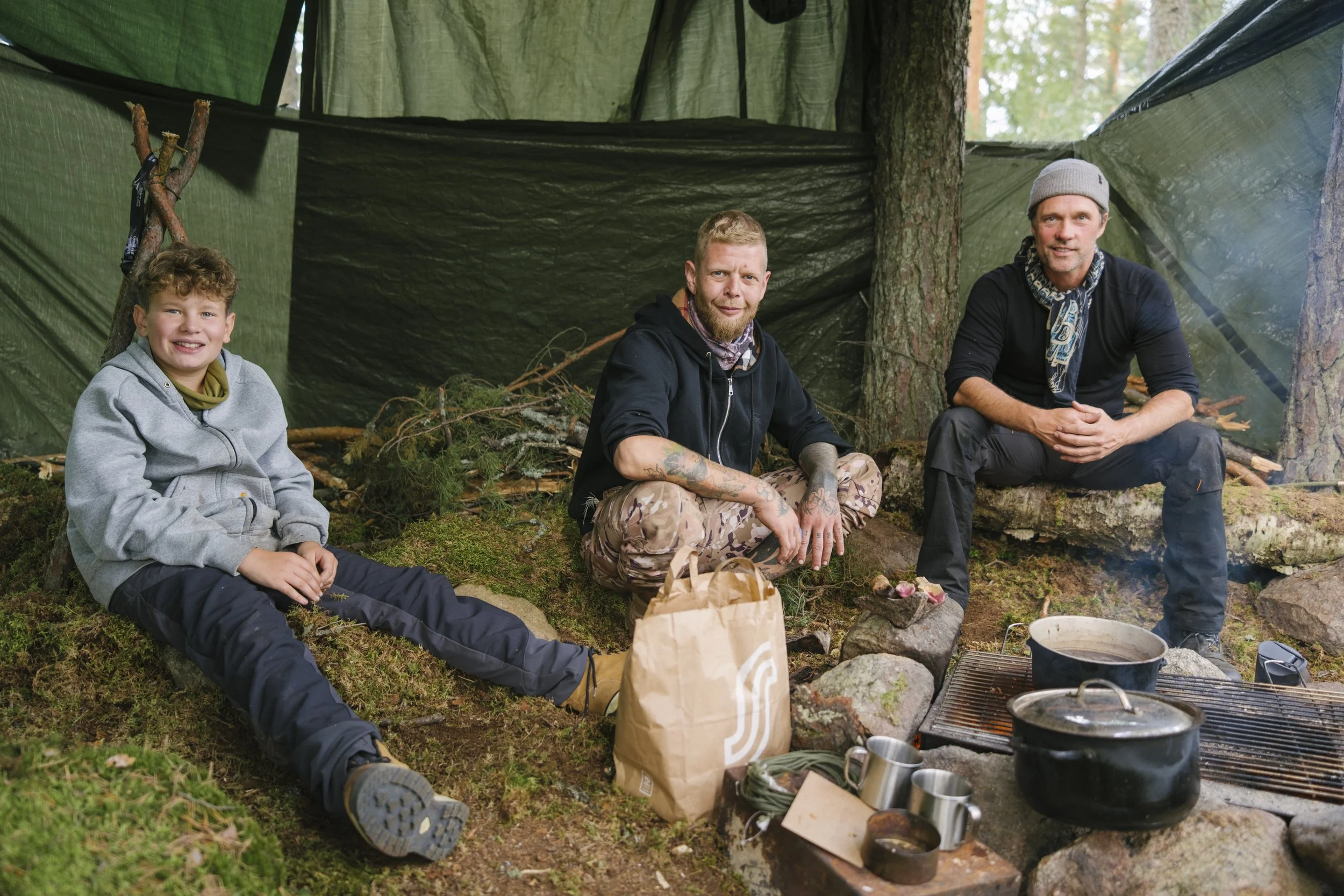 Drei Männer und ein Junge sitzen im Wald vor einem Lagerfeuer, umgeben von Campingausrüstung und Kochtöpfen.