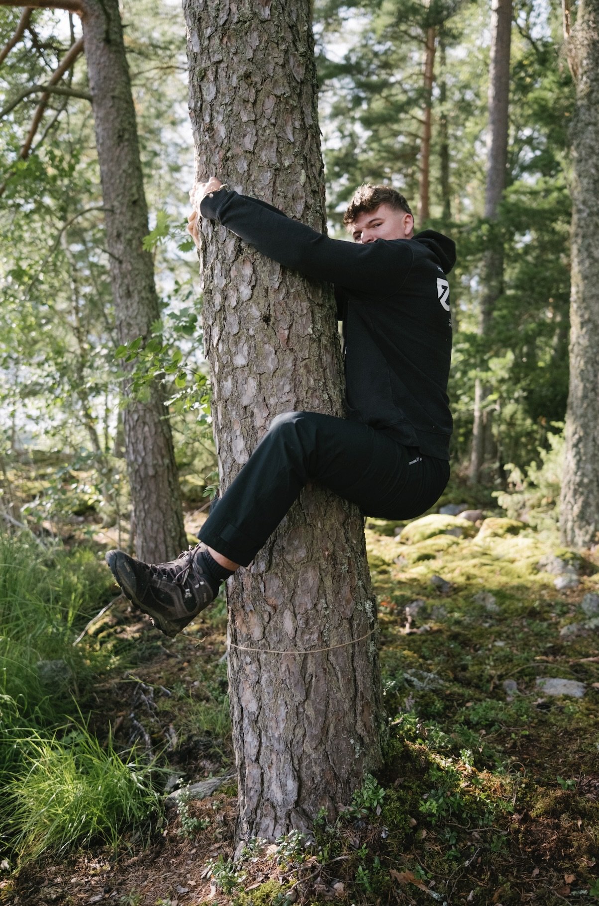 Junge Person zeigt Klettertechnik an einem Baum in einem Wald, trägt schwarze Kleidung und Wanderschuhe.
