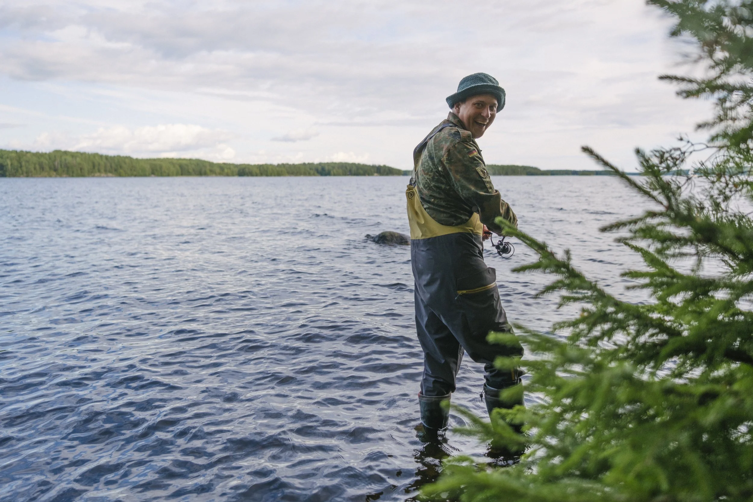 Ein lachender Mann beim Angeln im See, umgeben von Natur, im Frühjahr oder Sommer, trägt Regenbekleidung und einen Hut.