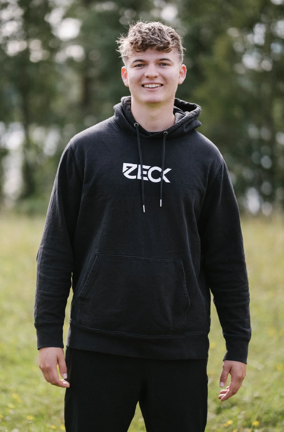 Ein junger Mann steht im Freien auf einer Wiese, trägt einen schwarzen Hoodie mit der Aufschrift 'ZECK' und lächelt in die Kamera.