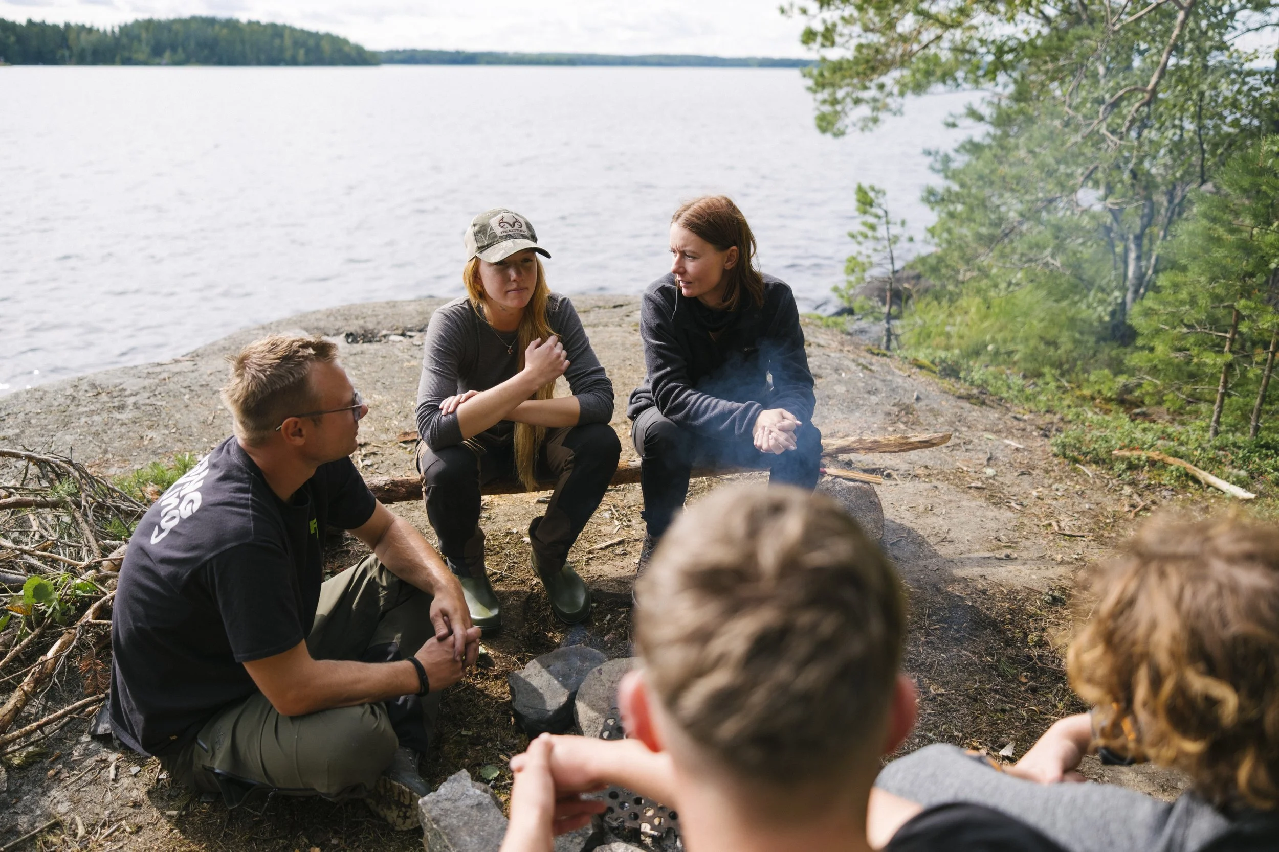 Gruppe von jungen Menschen am Lagerfeuer am See, im Gespräch, umgeben von Bäumen.