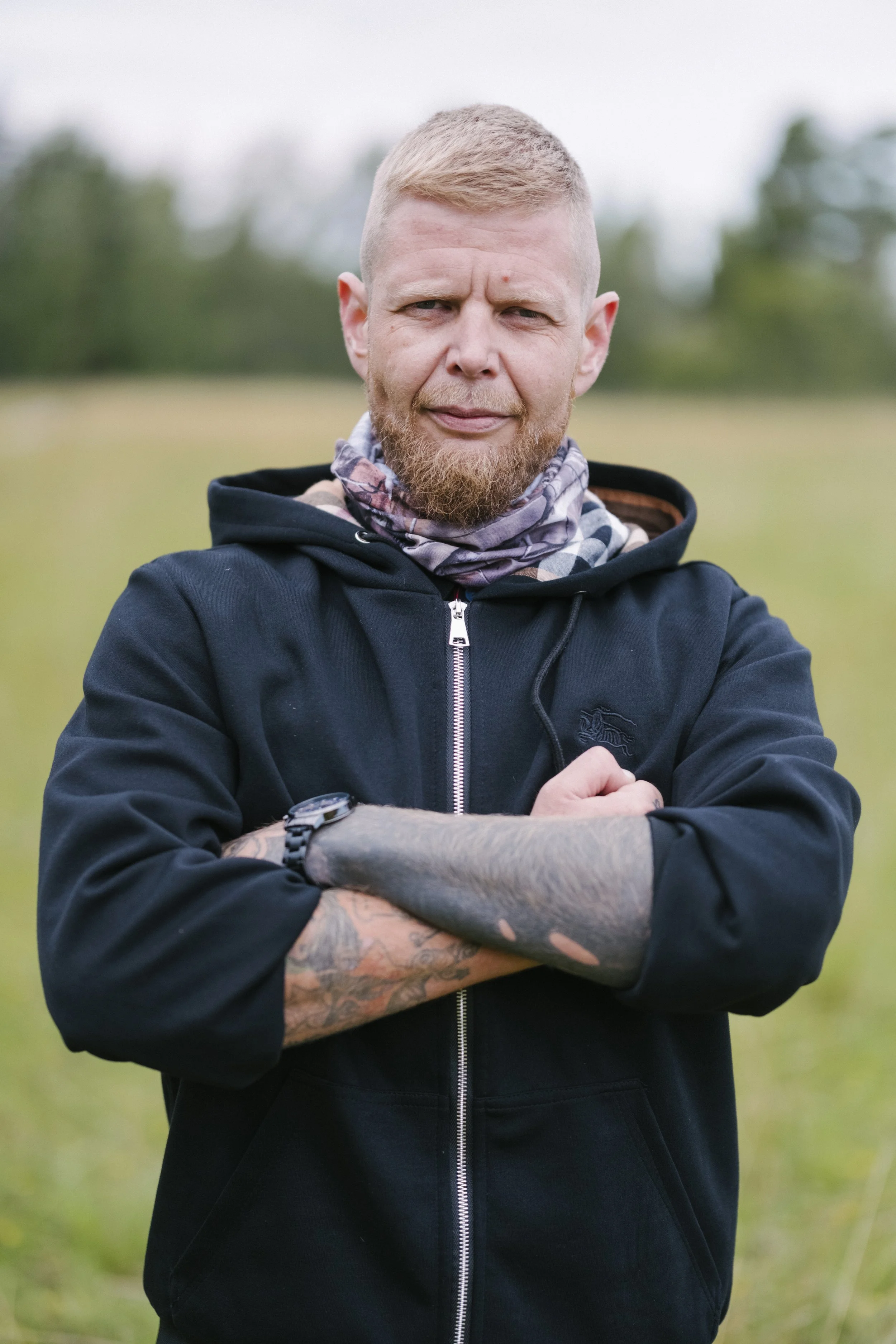 Mann mit kurzen blondem Haar und Bart, trägt schwarze Jacke und Armband, steht mit verschränkten Armen im Freien auf einer grünen Wiese.
