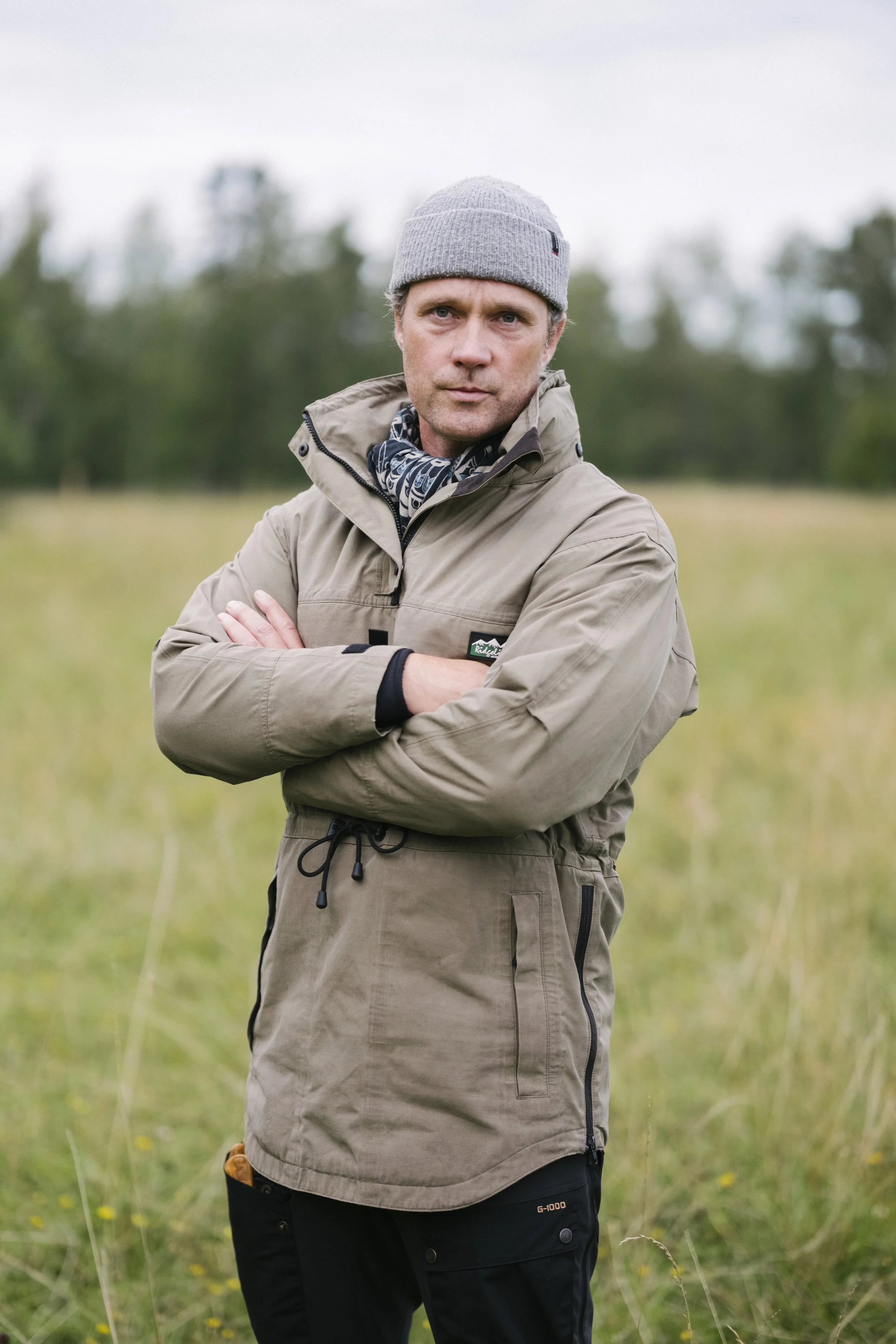 Mann steht mit verschränkten Armen in einem Grasefeld, trägt eine graue Mütze, eine beige Jacke und blickt ernst. Im Hintergrund sind Bäume und der Himmel sichtbar.